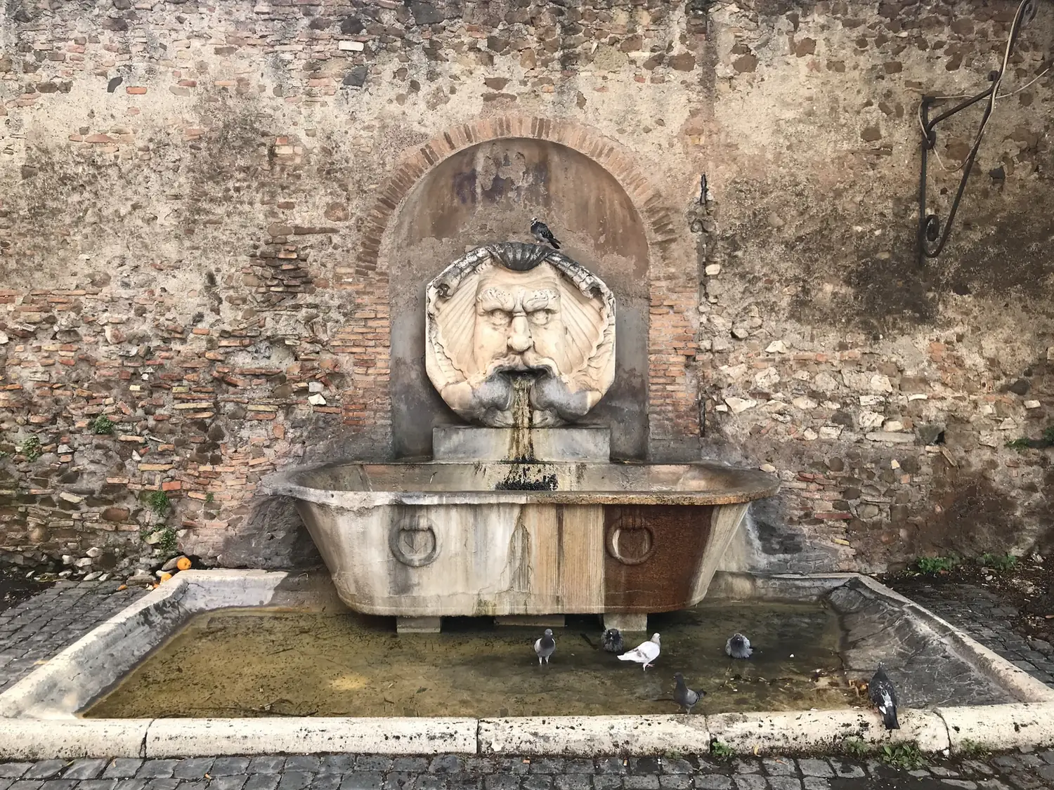 A carved stone mask fountain against a brick wall on the Aventine's Piazza Pietro d'Illiria.