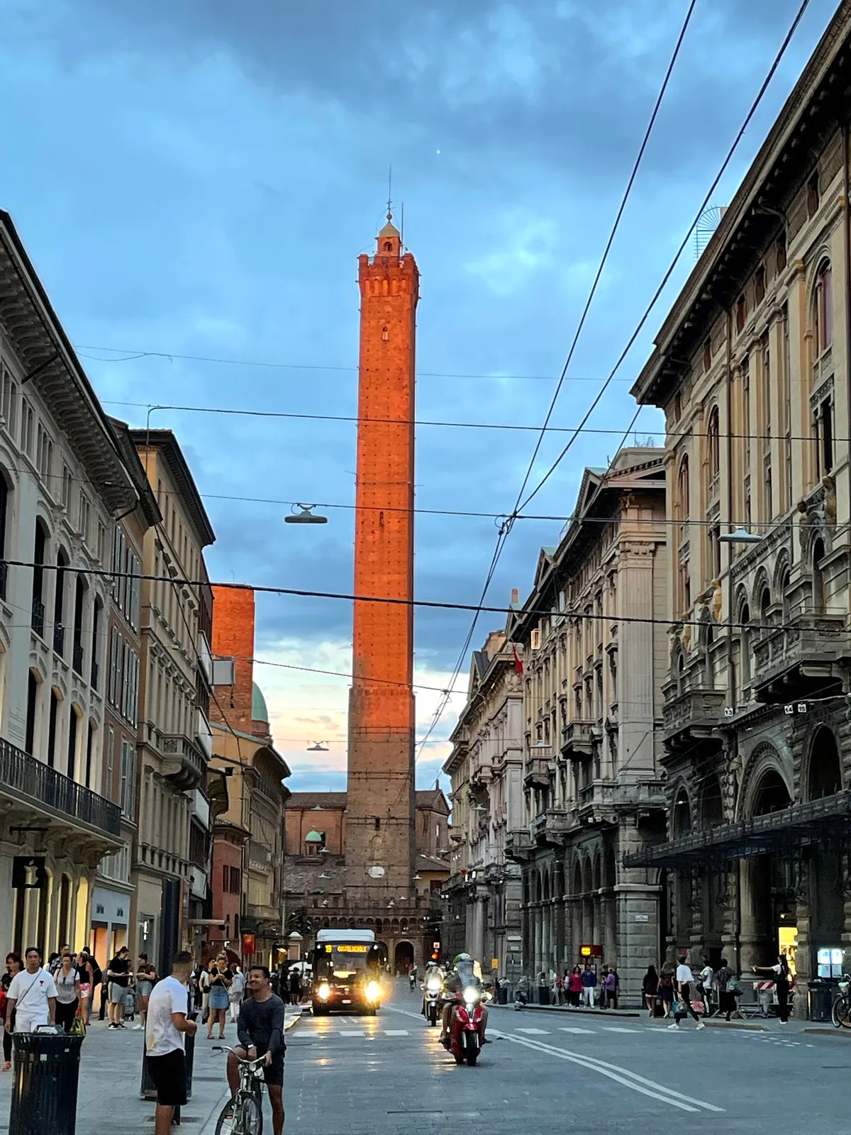 Via Rizzoli at dusk with people walking and the lit Asinelli tower ahead.