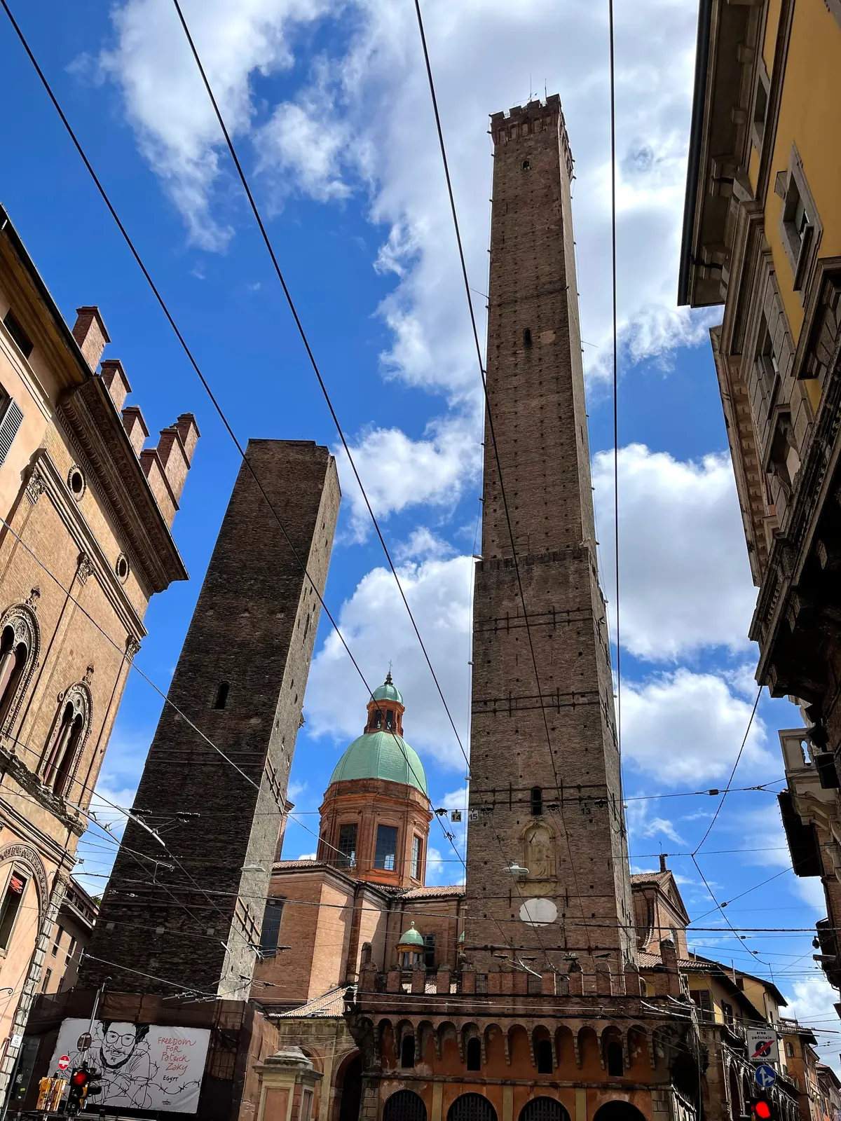 The two towers of Bologna above rooftops with a church dome under a blue sky.