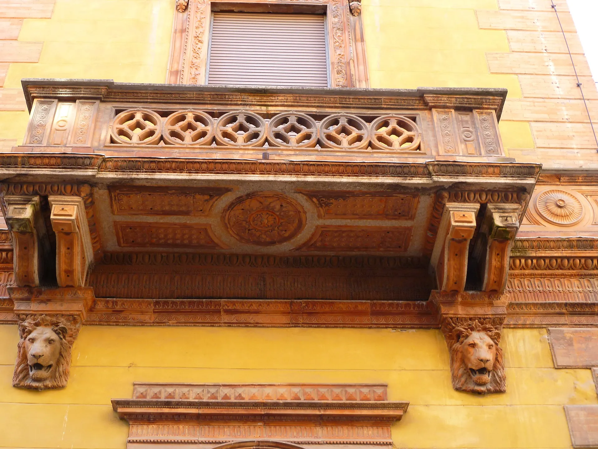 Close view of a carved stone balcony with lion-head brackets on a yellow palazzo wall in Ferrara.