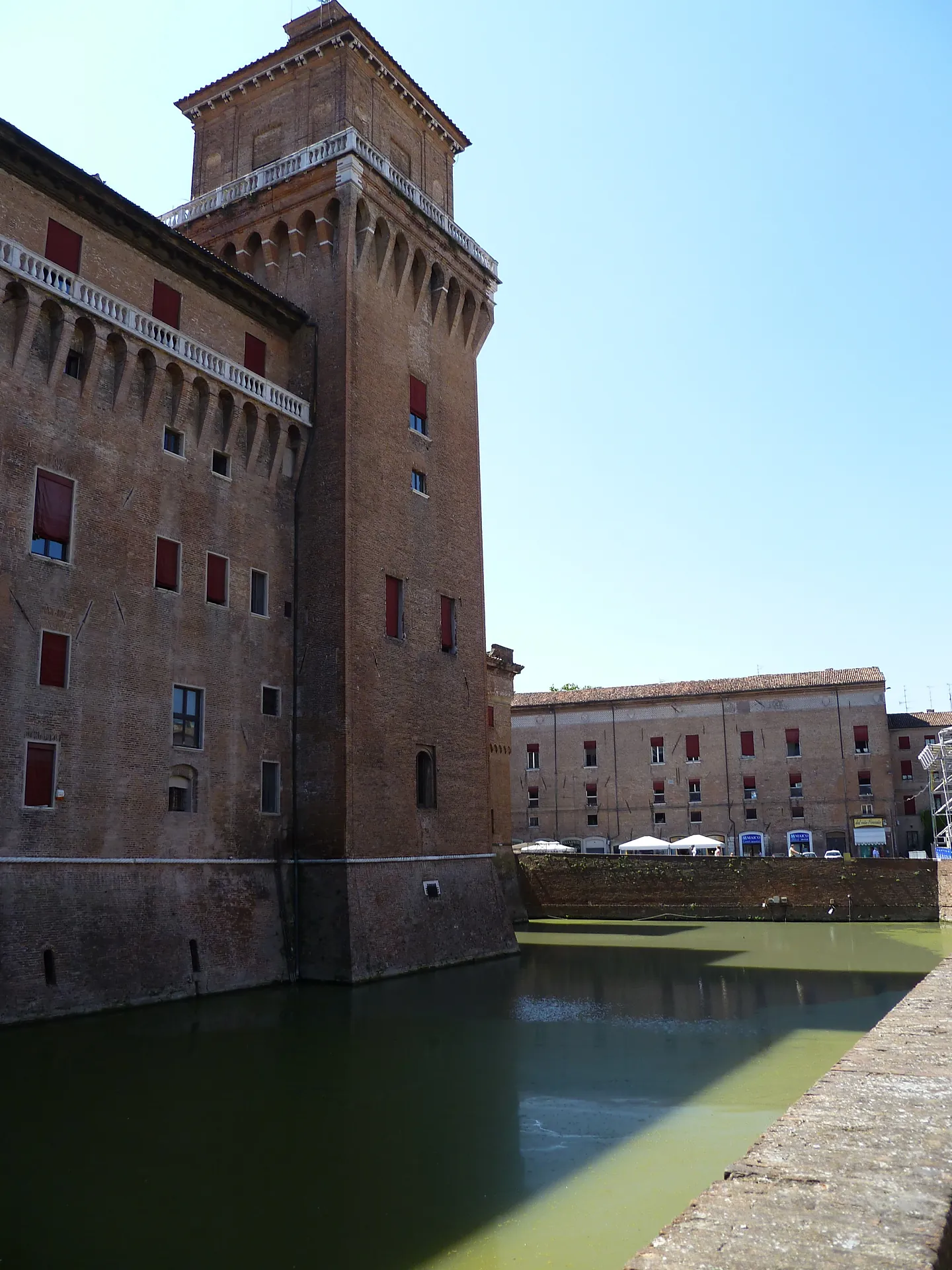 The brick Castello Estense in Ferrara reflected in its green moat under a clear sky.