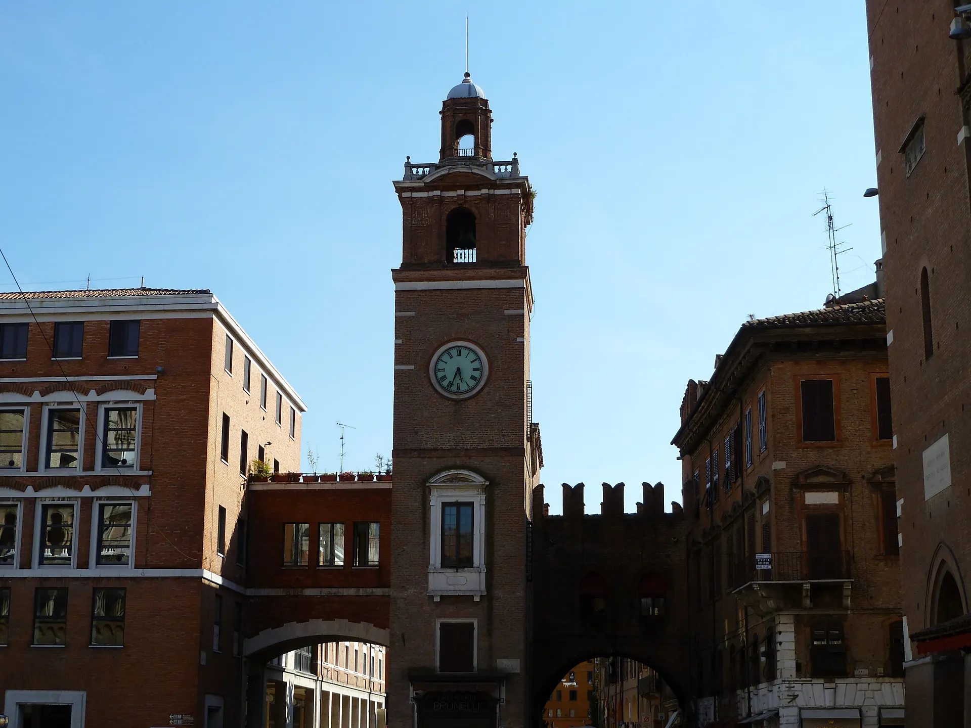 A tall brick clock tower with a white belfry rising between old buildings in central Ferrara.