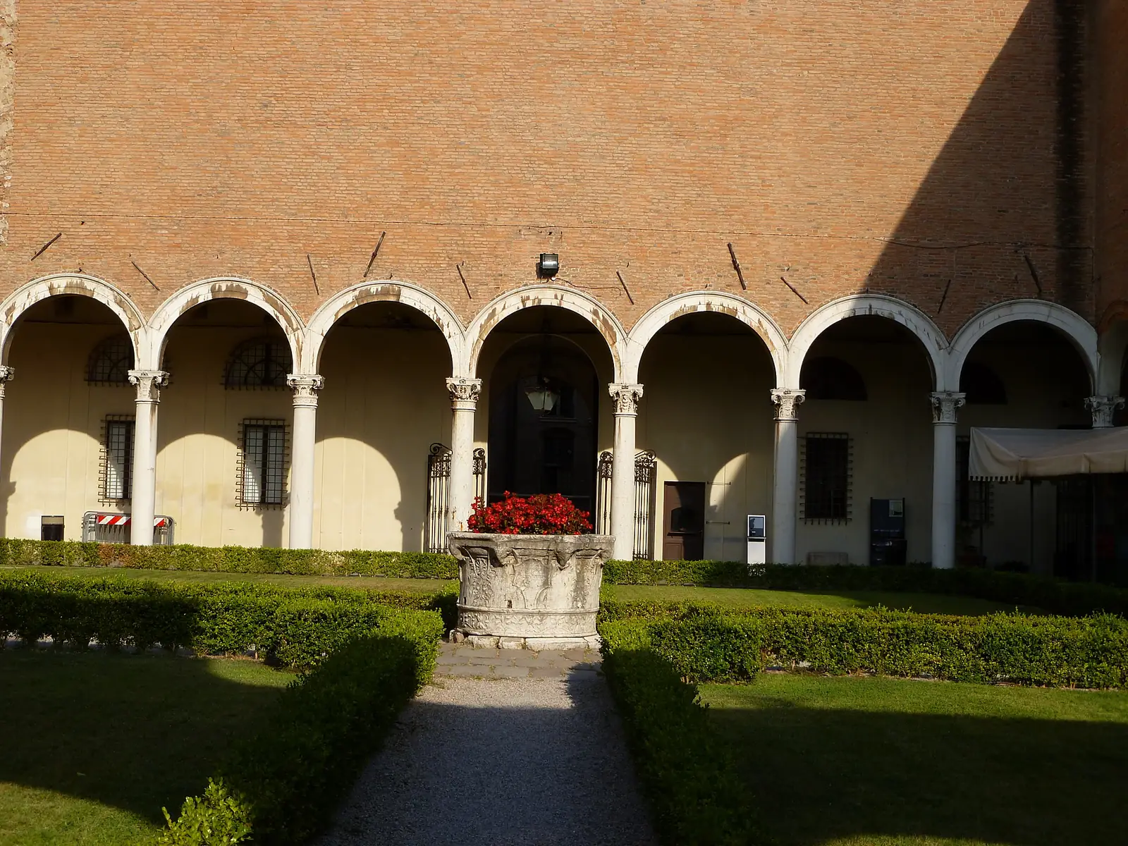 A Renaissance courtyard in Ferrara with a two-story arcaded loggia, clipped hedges, and a marble wellhead.