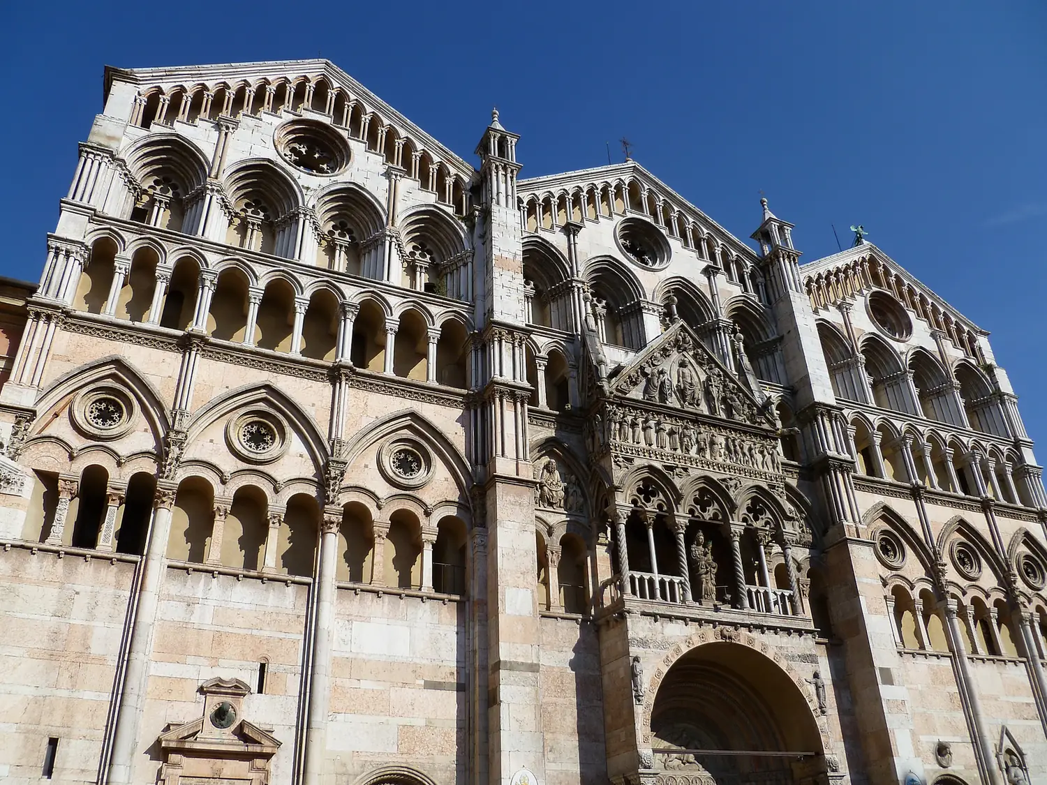 The white marble west front of Ferrara Cathedral with Romanesque arches below and Gothic arcaded gables above.