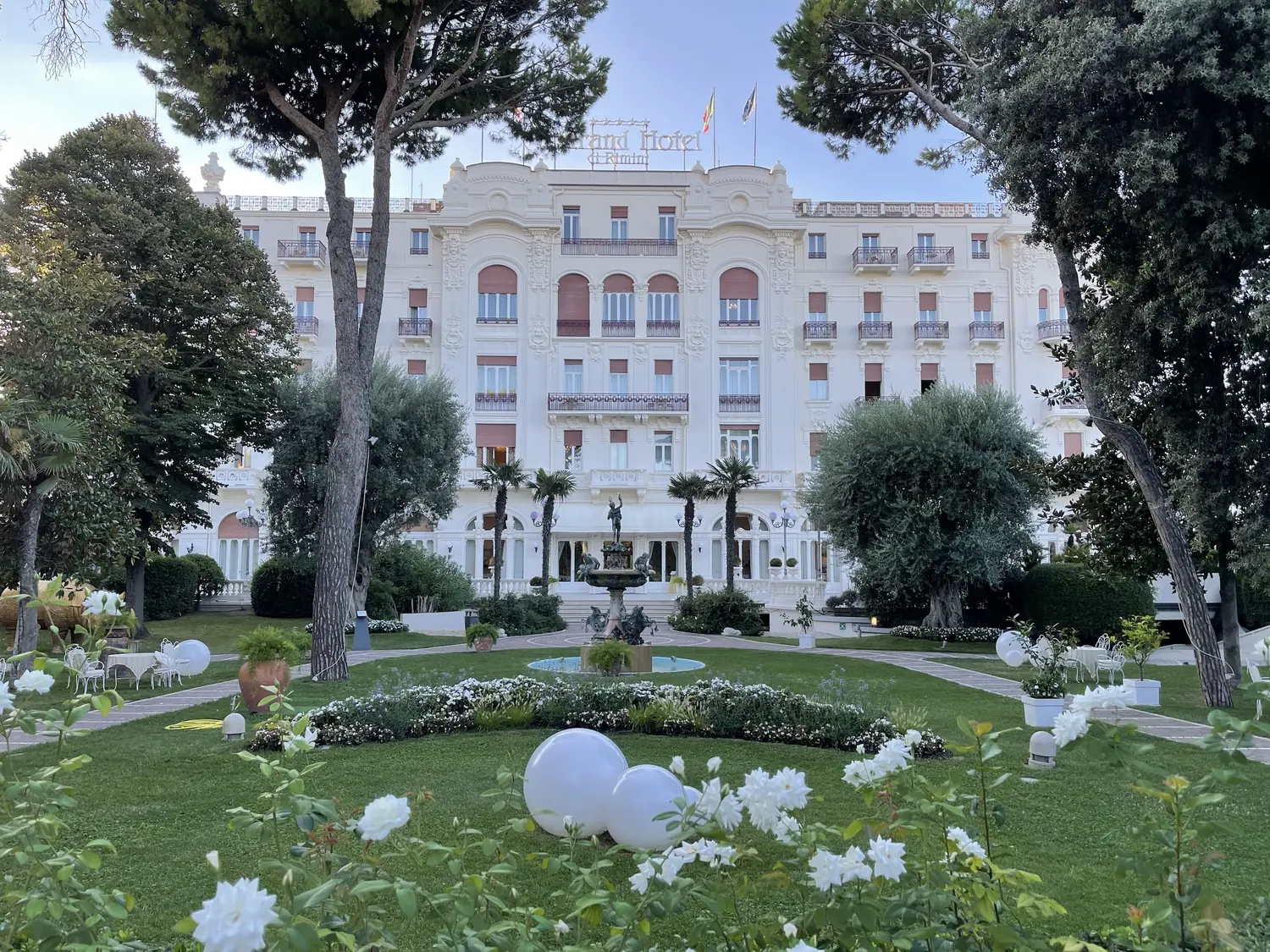 The white Liberty facade of the Grand Hotel Rimini seen across its garden and umbrella pines