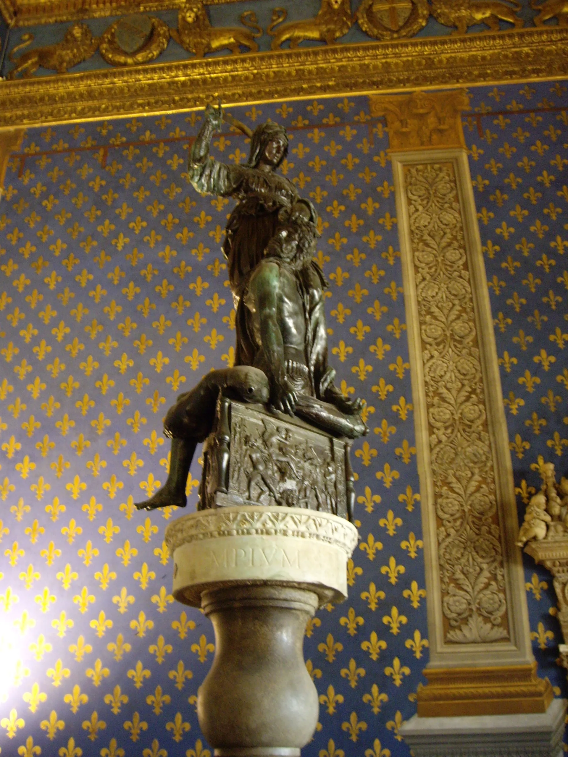 Donatello's bronze Judith and Holofernes standing against the blue and gold lilies of the Sala dei Gigli