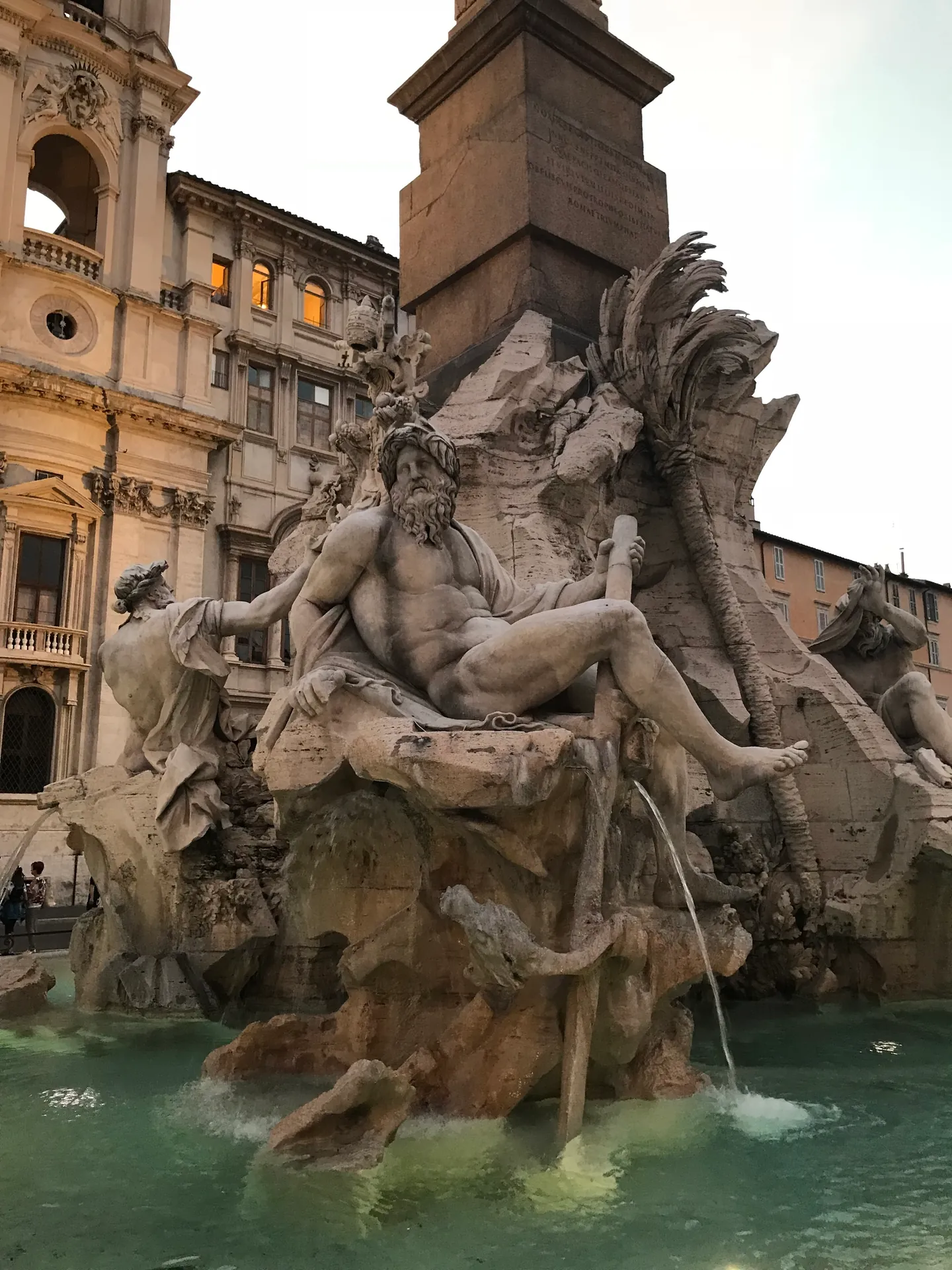 Ganges river god statue at dusk with lit palace windows behind at Piazza Navona