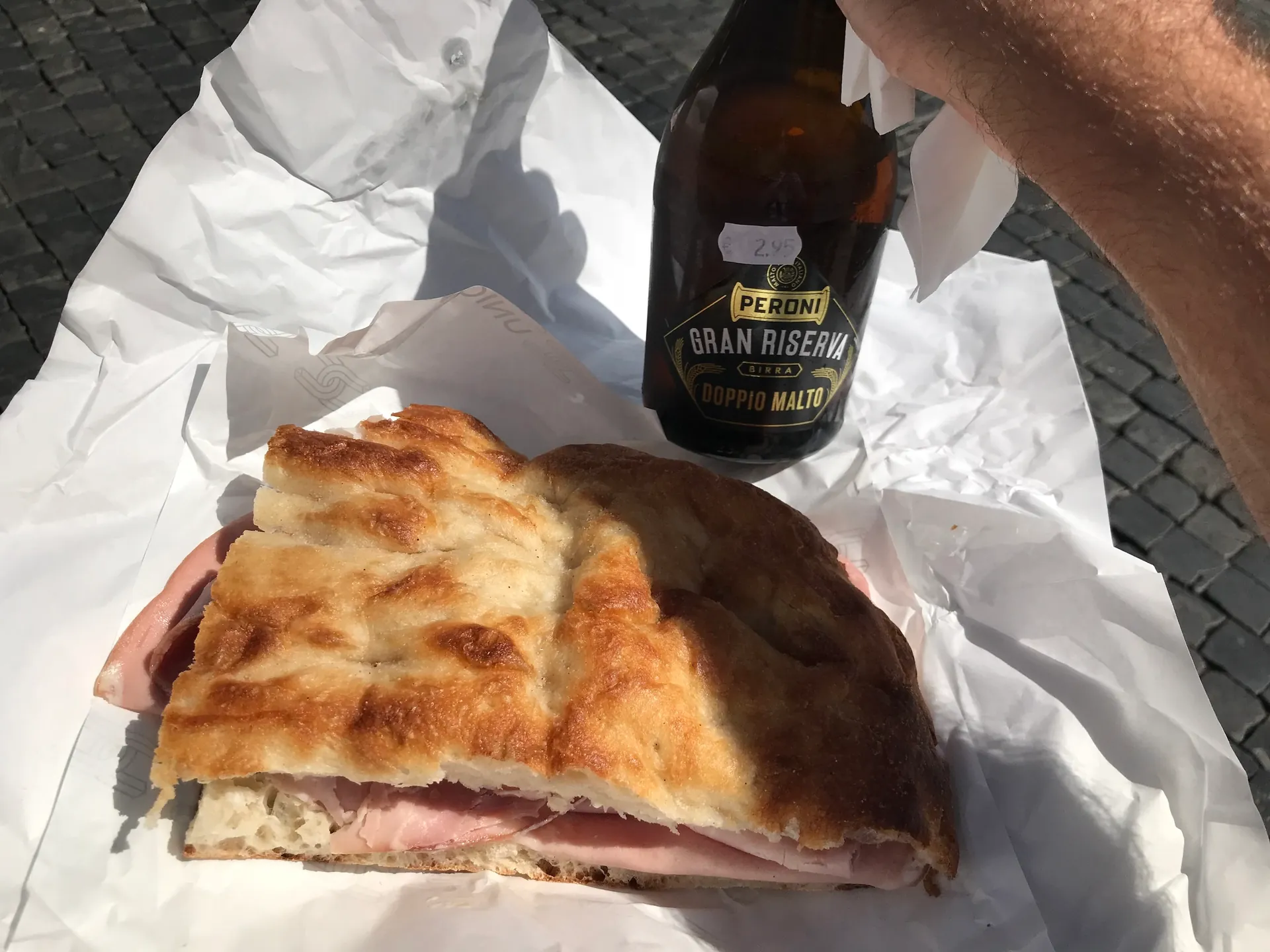 Focaccia sandwich and a bottle of Peroni Gran Riserva held above the cobblestones of Piazza Navona