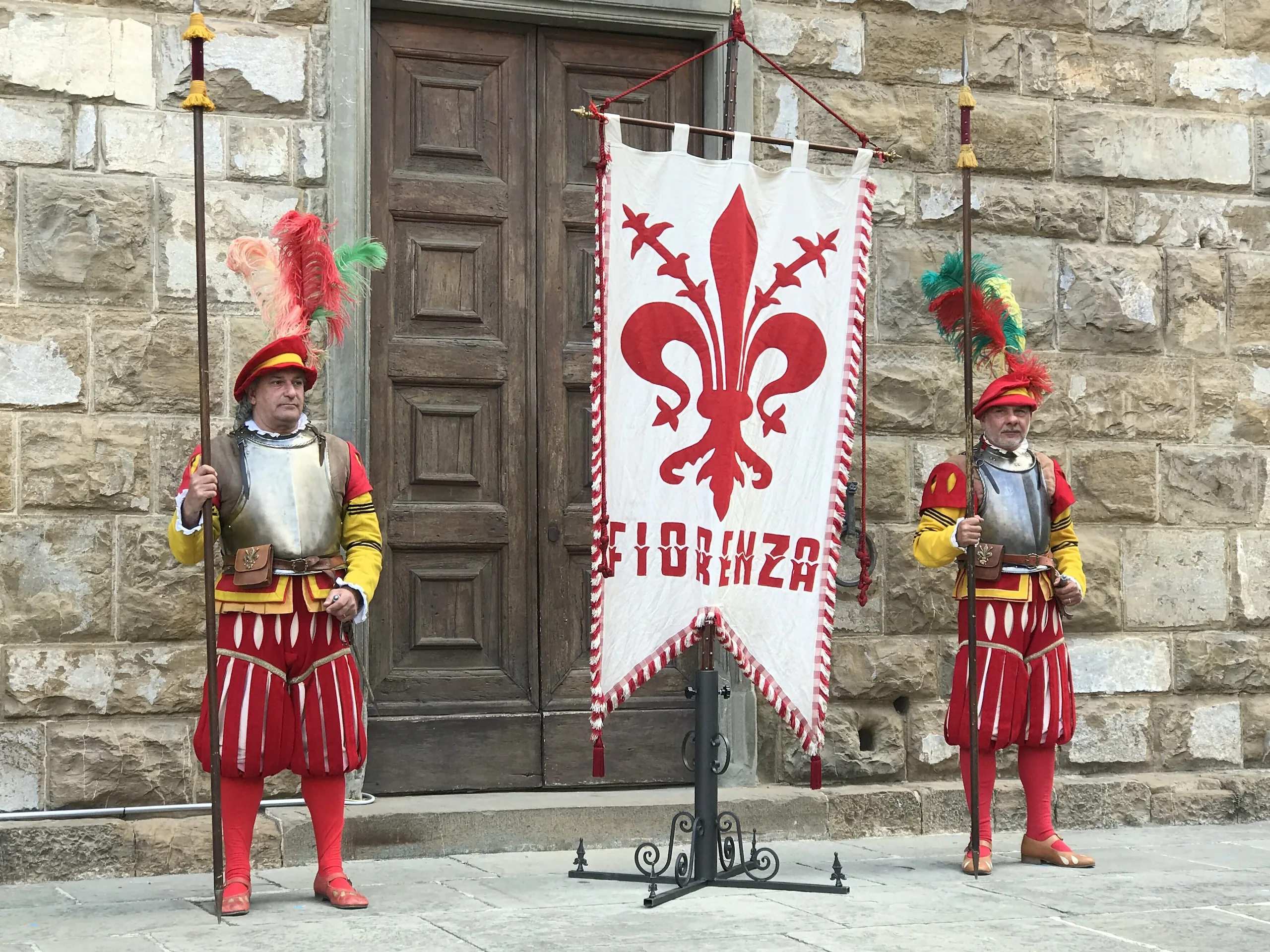 Two guards in red and yellow Renaissance livery with plumed helmets flanking the white banner of Florence with its red lily