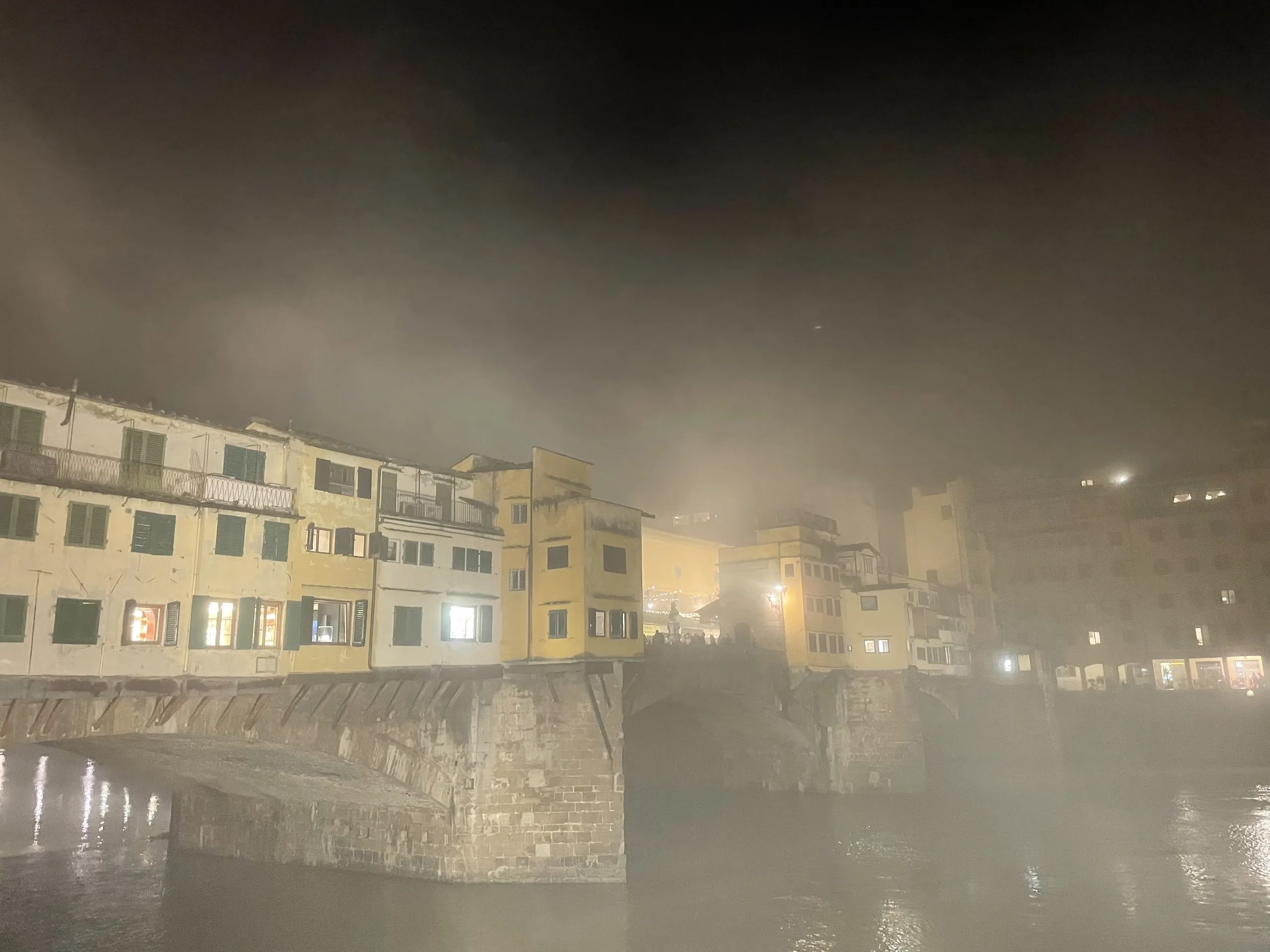 The Ponte Vecchio at night with fog rolling off the Arno, its shop windows lit through the haze