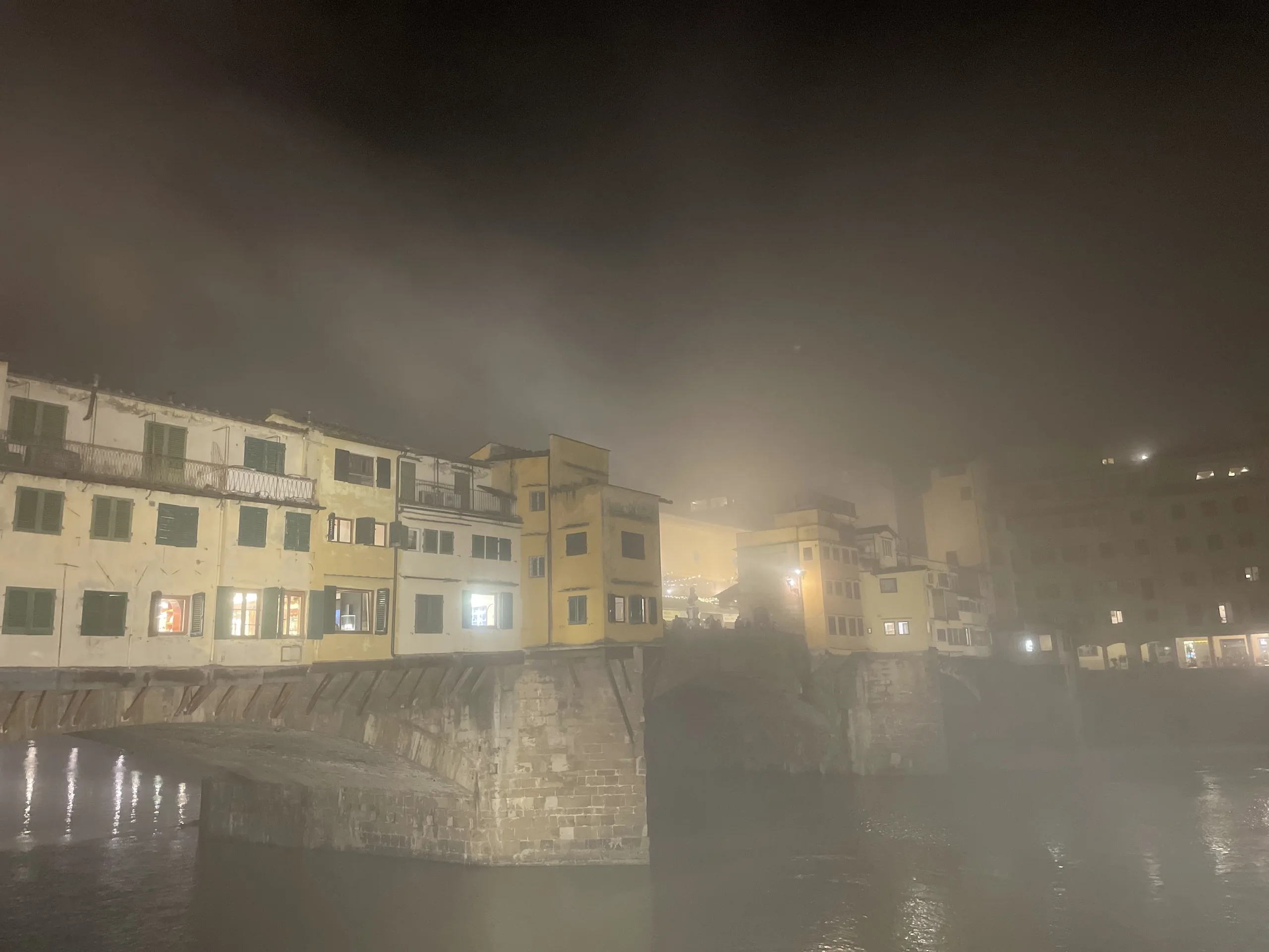 The lit windows and shuttered fronts of the Ponte Vecchio glowing through river fog at night