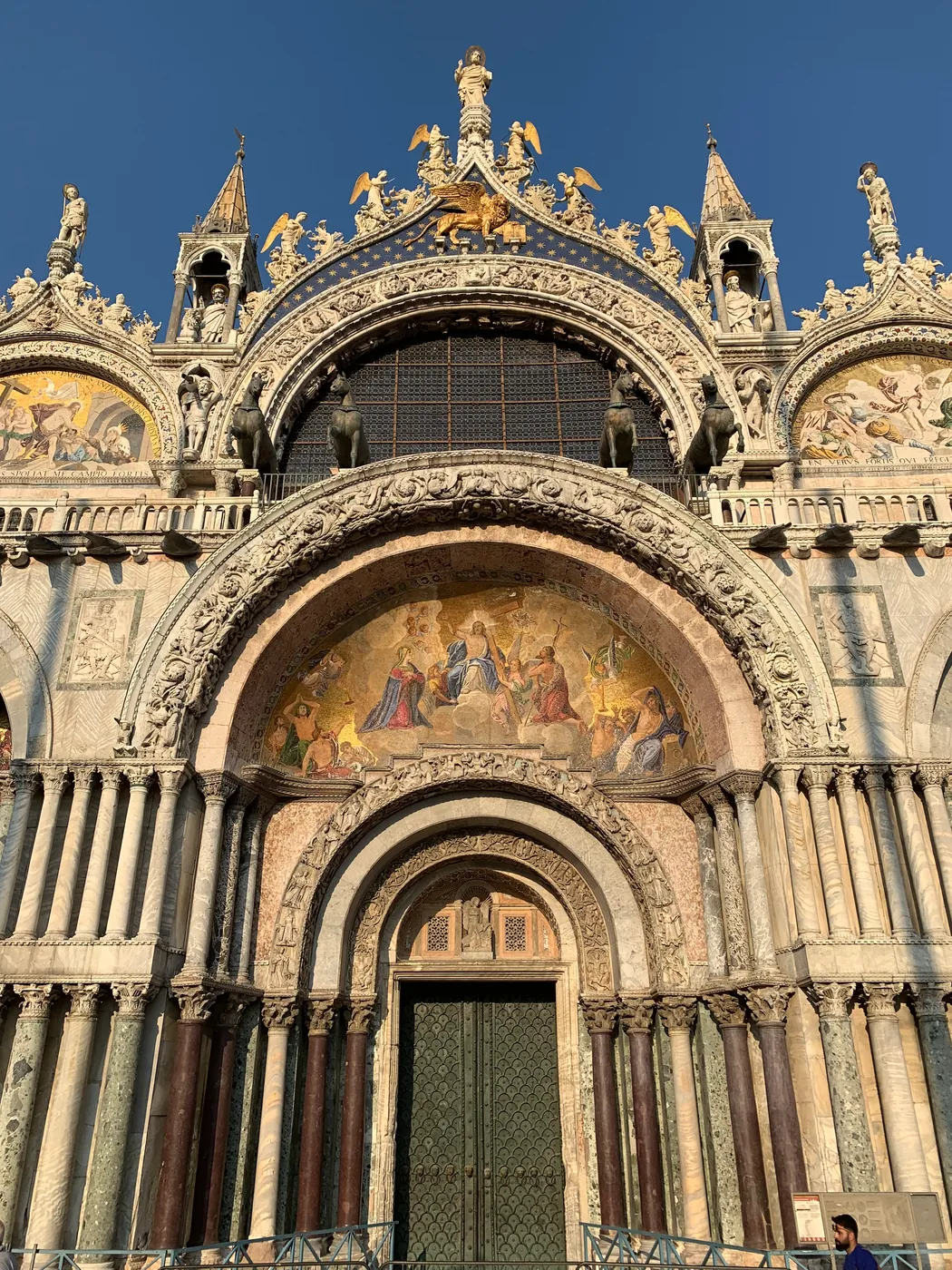 Upper facade of St Mark's Basilica with gilded mosaics, ogee arches and statues against a blue sky