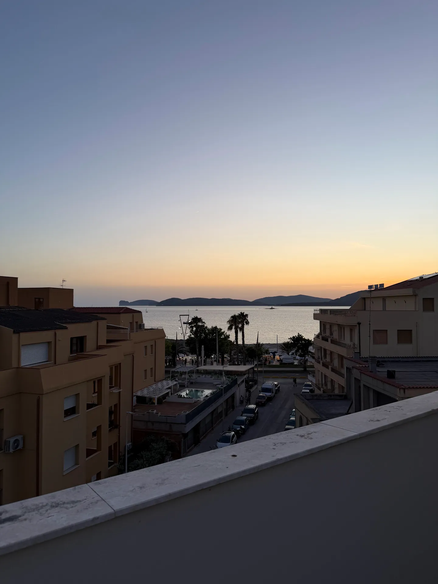 Rooftops and a street leading to a bay at dusk, amber sky behind a distant headland.