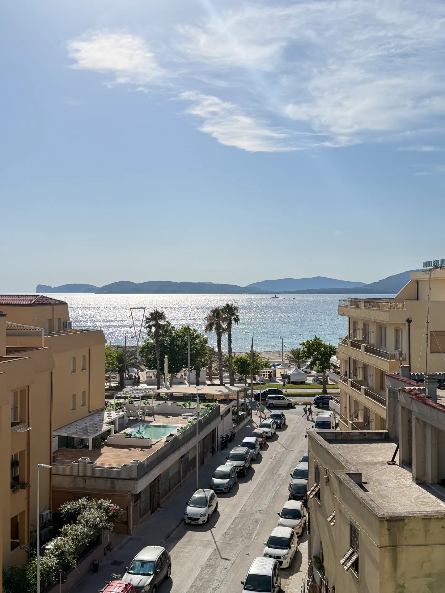 A street running down between buildings to palms and a sunlit bay with a headland beyond.