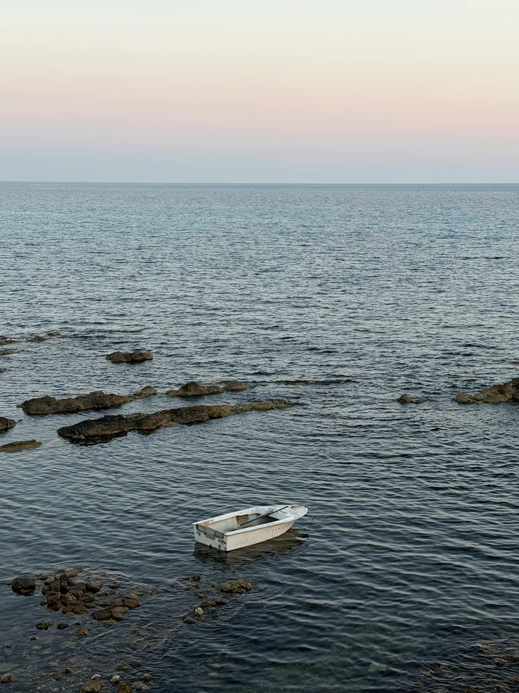 A small white rowing boat on calm water among low rocks at dusk below the sea walls.
