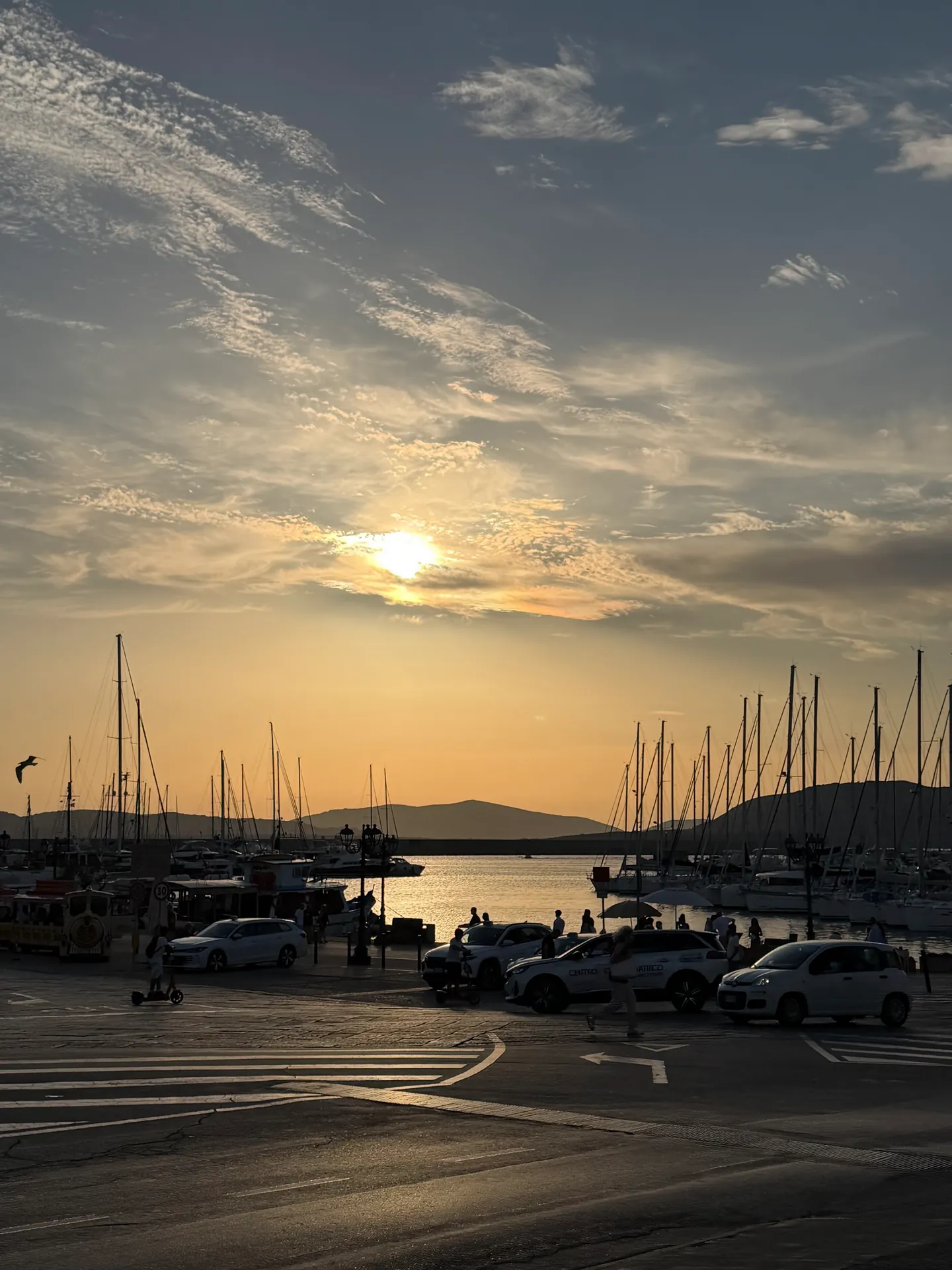 Sailing boat masts in Alghero's marina at sunset, hills silhouetted across the water.