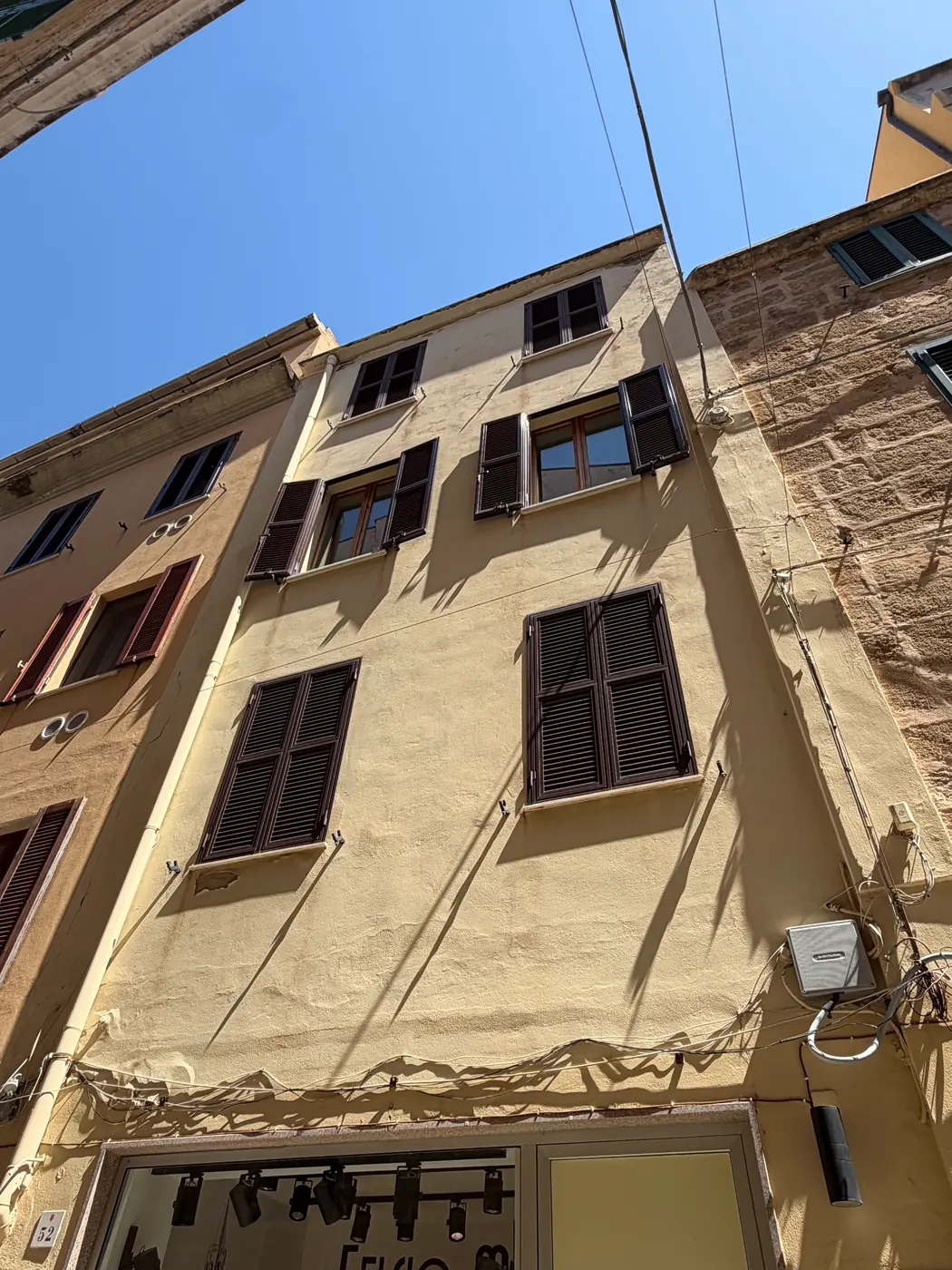 A yellow stuccoed facade with dark shutters seen from below in a narrow Alghero street.