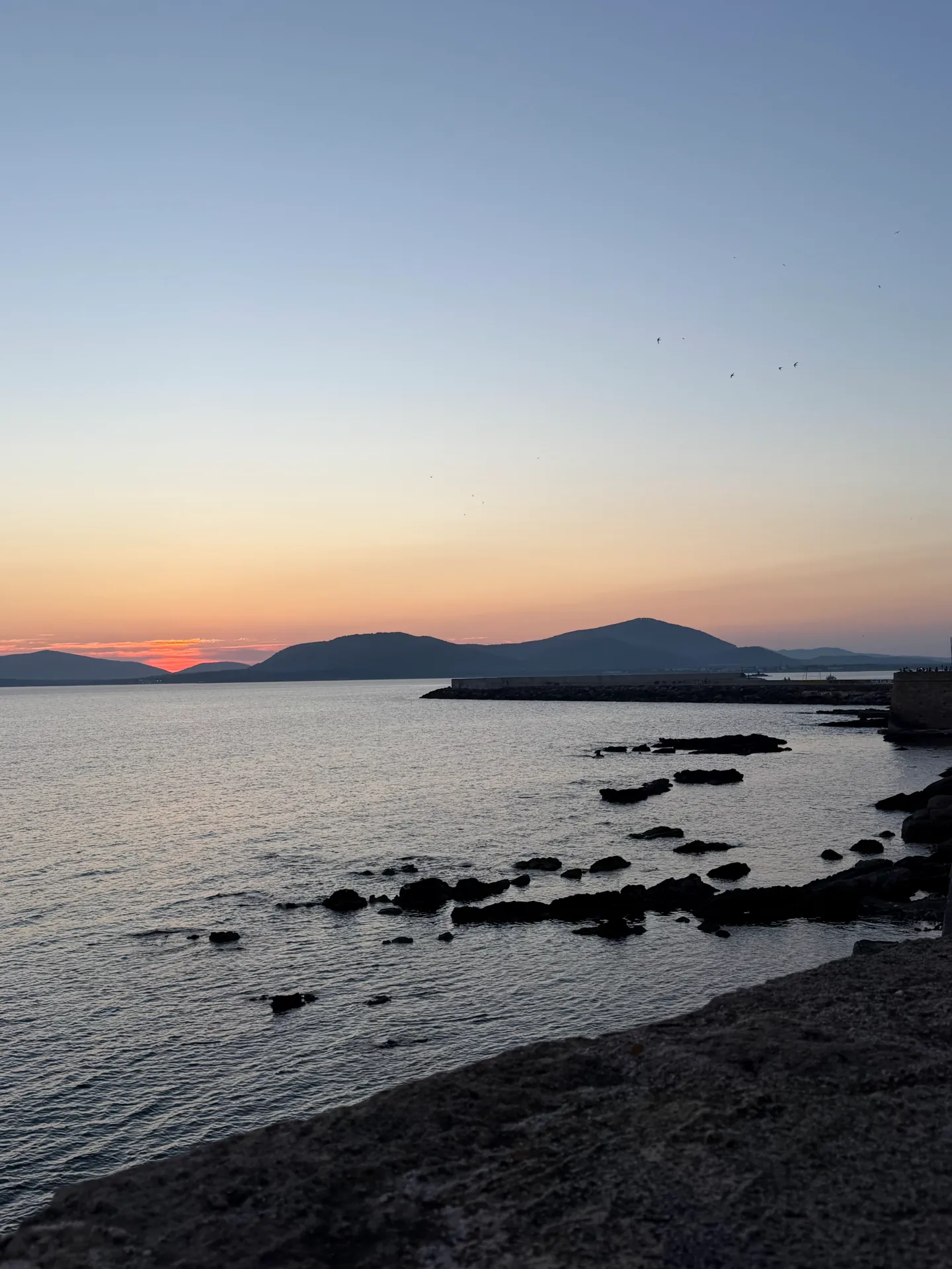 The sun setting behind low hills across a bay, seen from Alghero's ramparts over rocks.