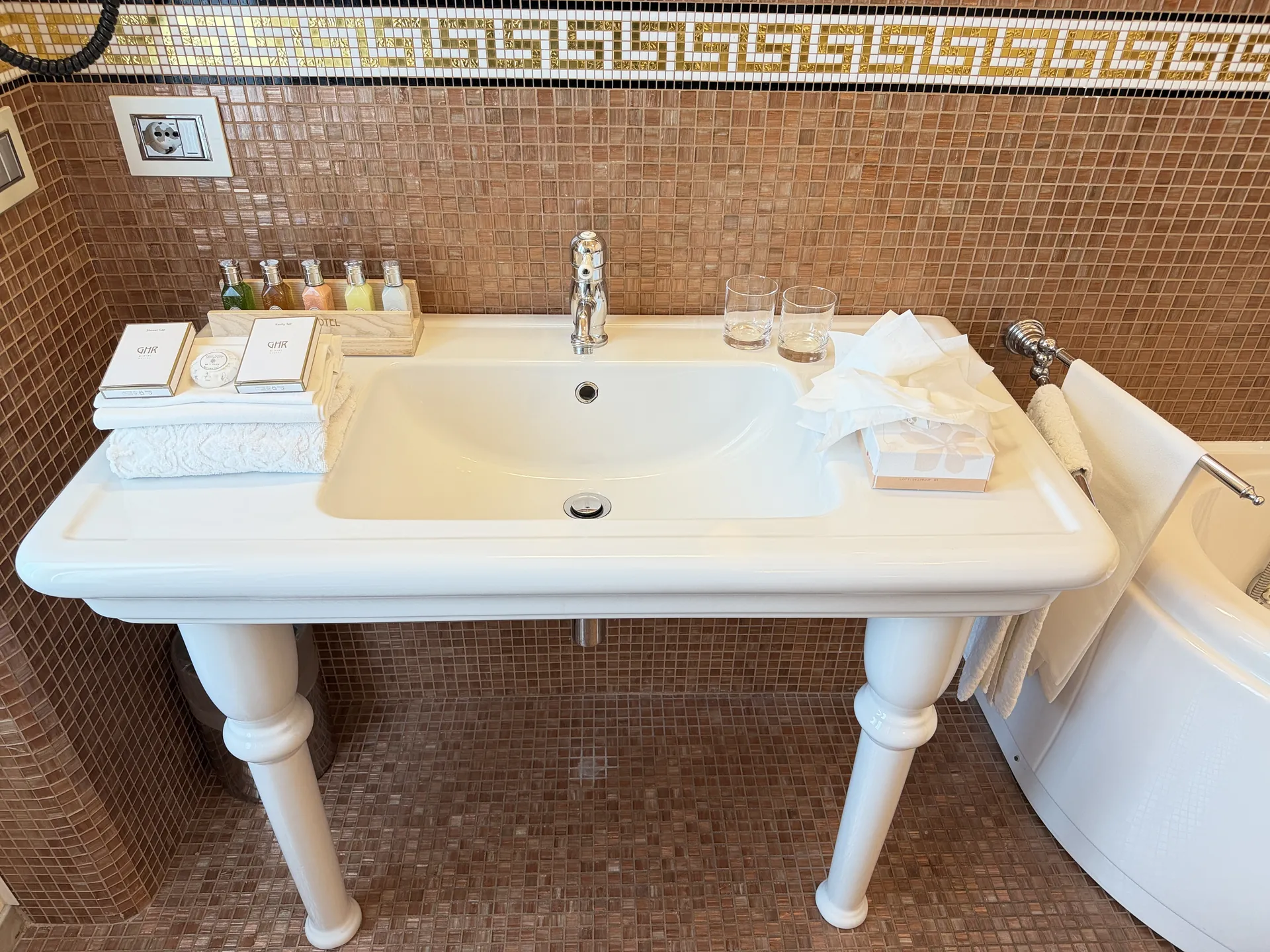 Twin basin console on turned legs against brown mosaic tiling with a Greek key border