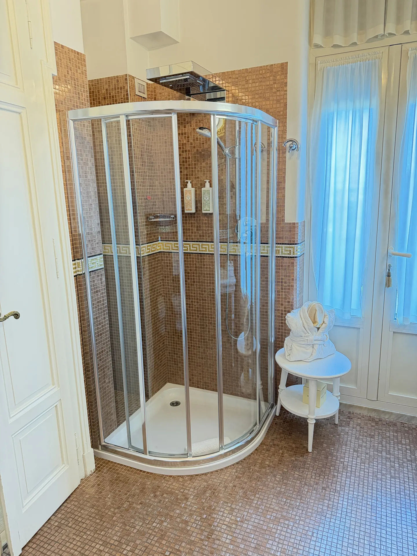 Quadrant glass shower beside a shuttered window in a mosaic-tiled hotel bathroom