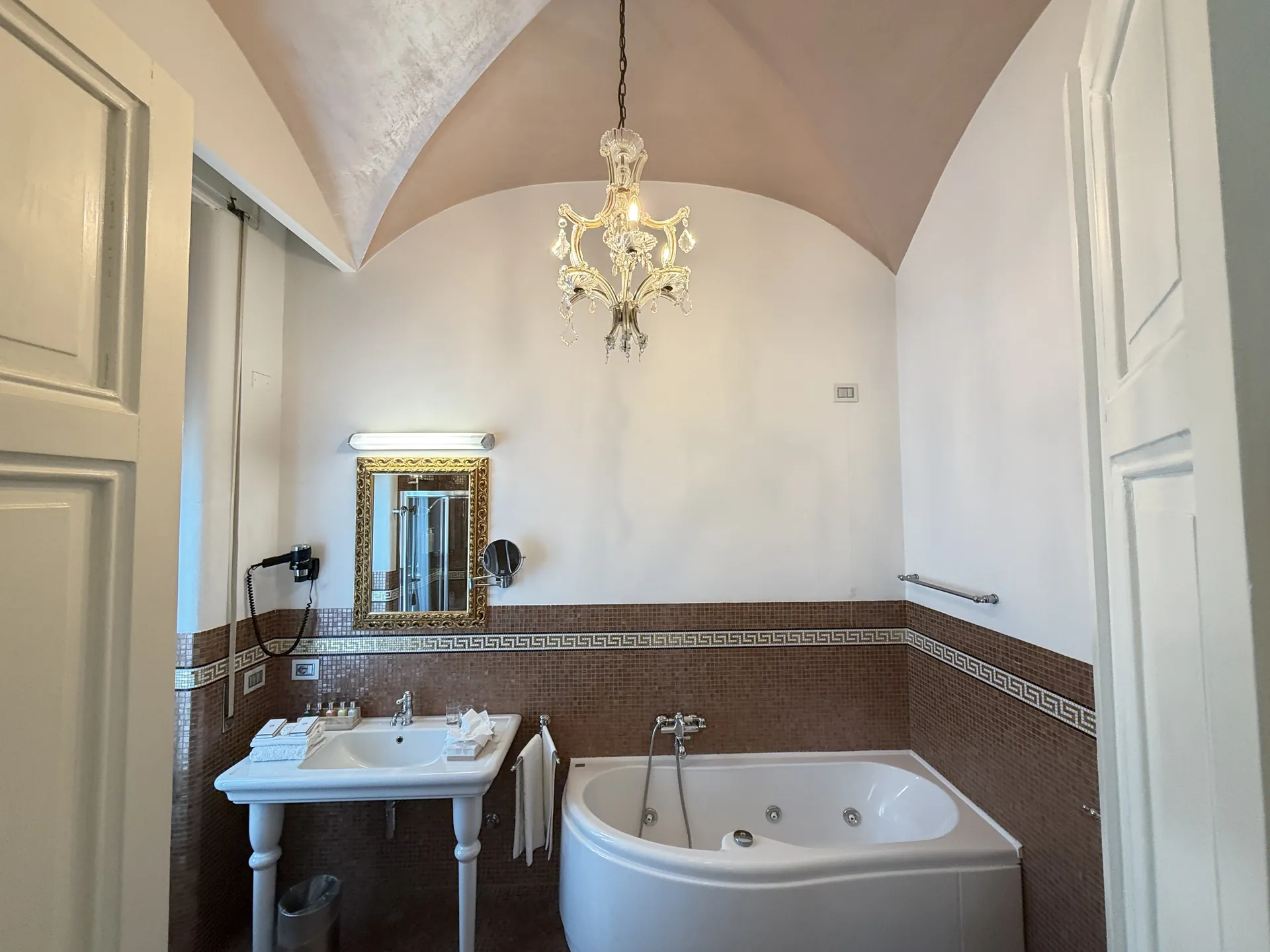 Barrel-vaulted hotel bathroom with a corner tub, mosaic tiling and a crystal chandelier