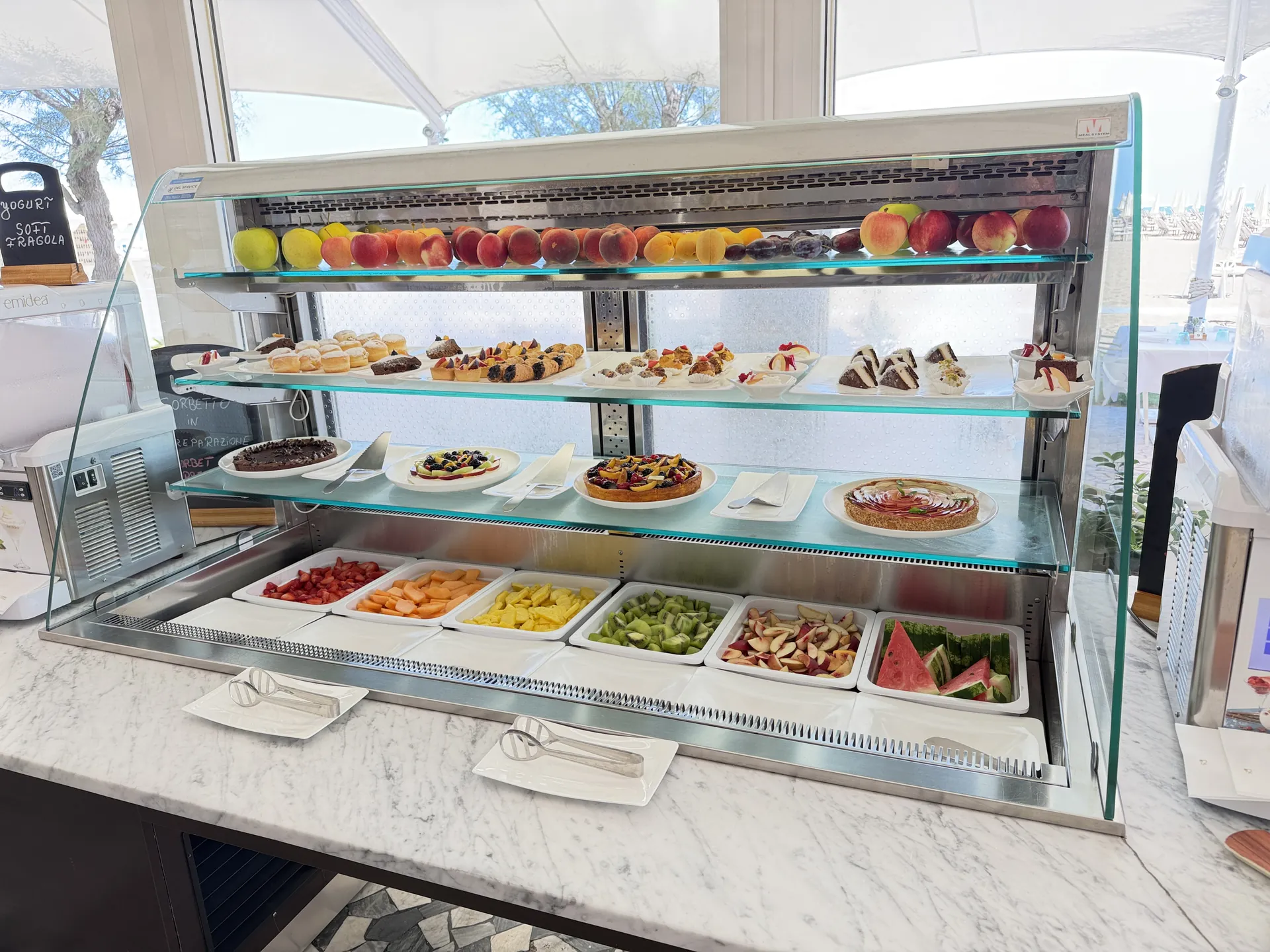 Glass case of tarts and pastries above trays of cut fruit at the beach buffet