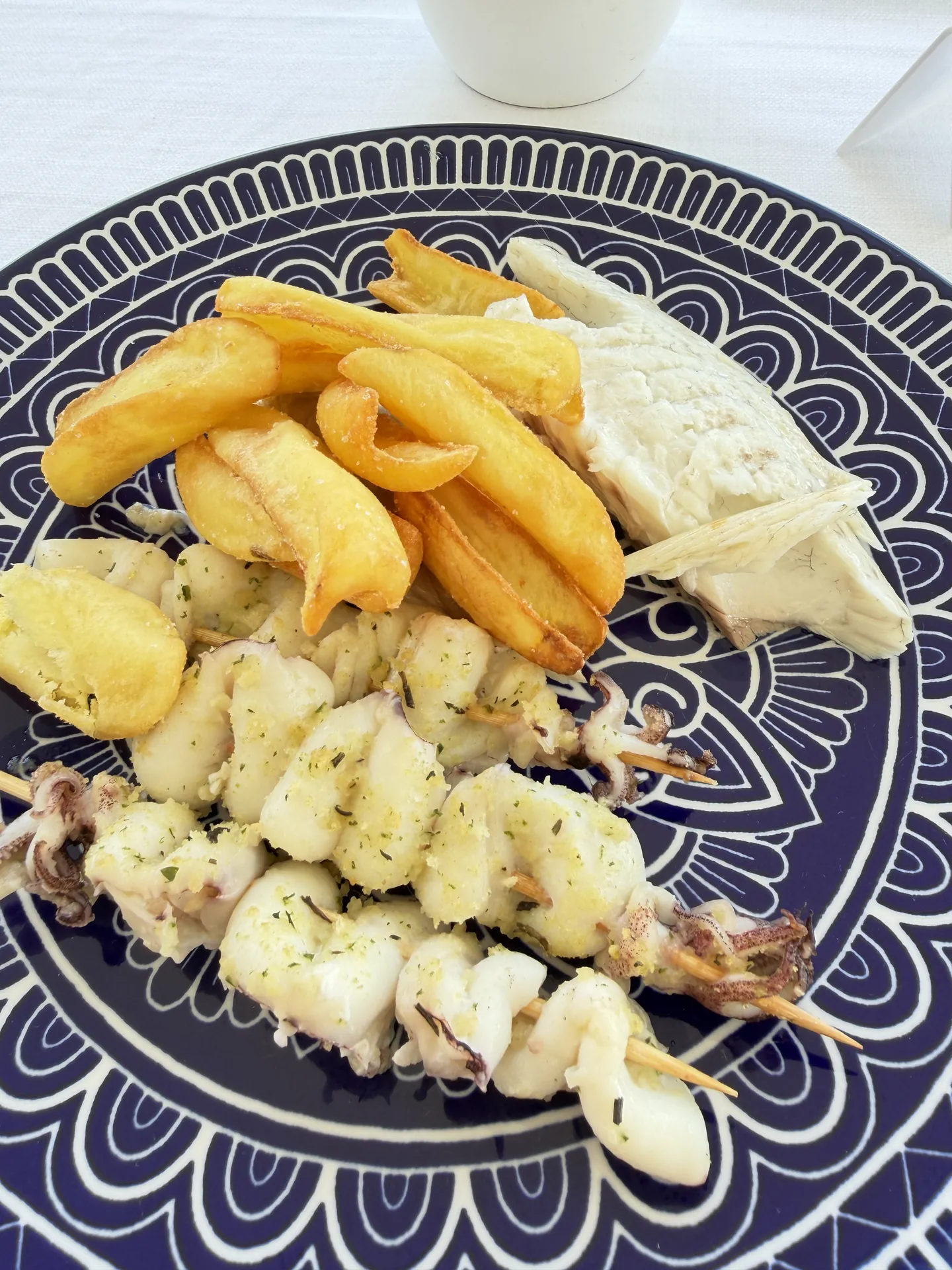 Squid skewers, fried potatoes and white fish on a blue and white plate