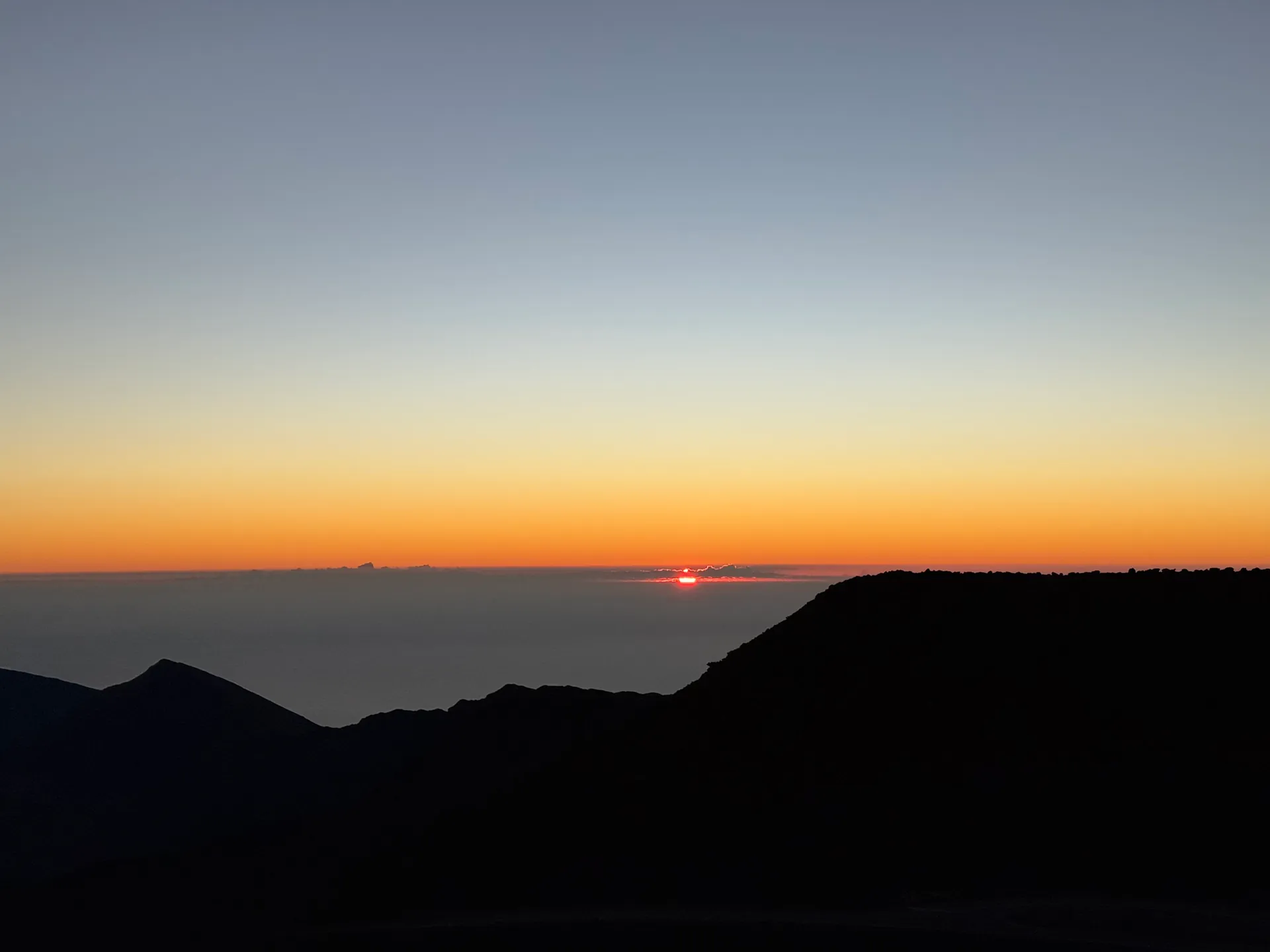 First sliver of sun on the horizon above a cloud deck with a cinder cone silhouette