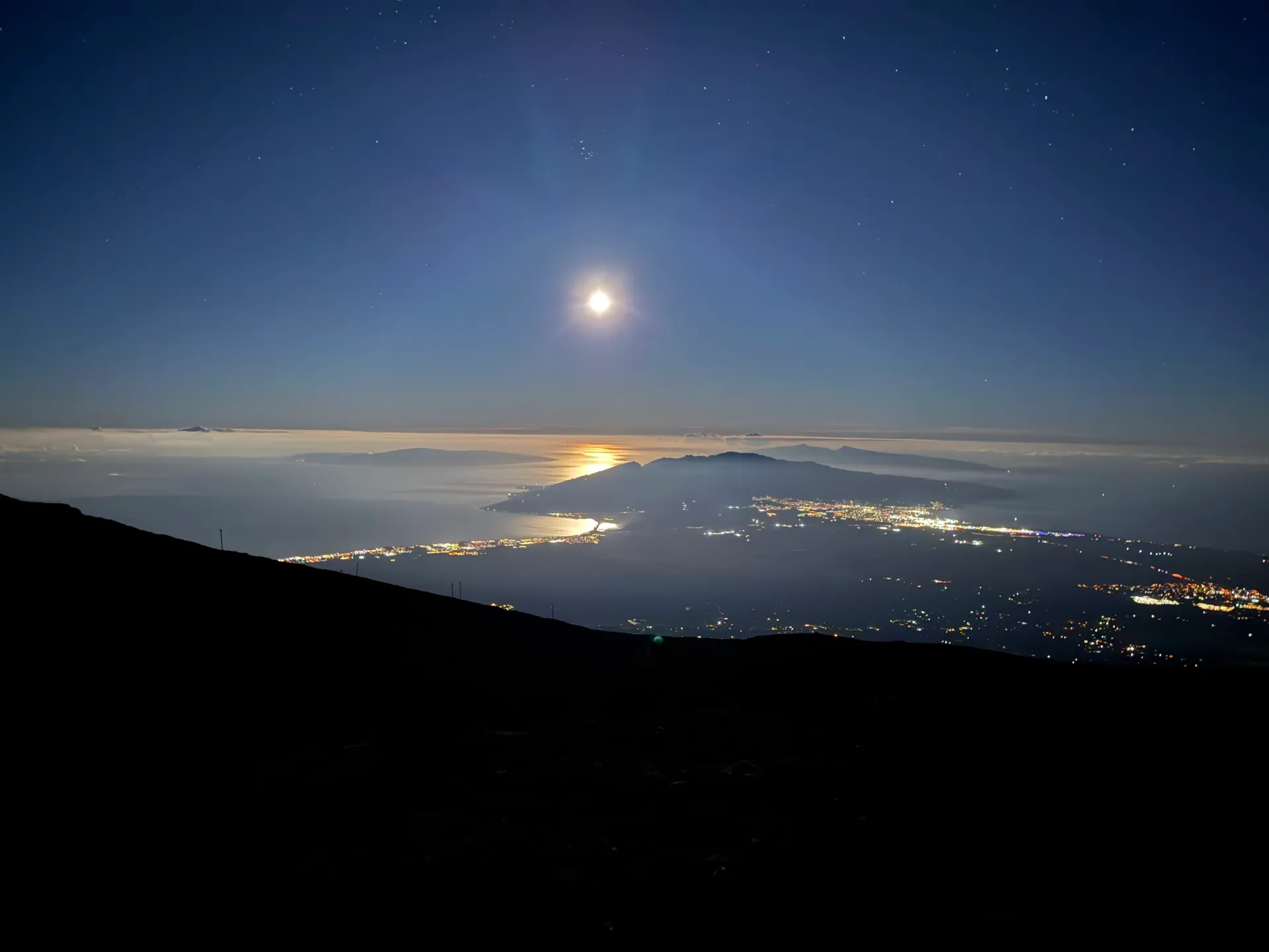 Moon over the ocean before dawn with coastal town lights below a cloud layer
