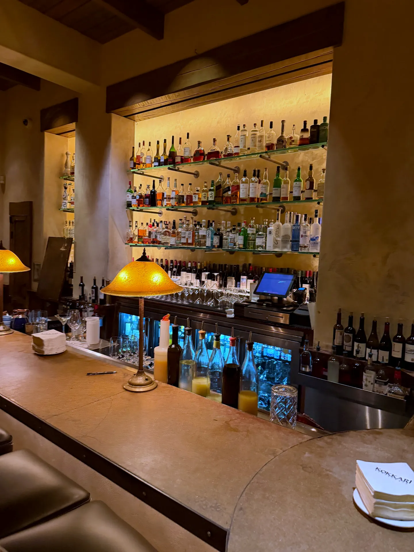 The long bar at Kokkari with a lit backbar of bottles and shaded lamps along the counter.