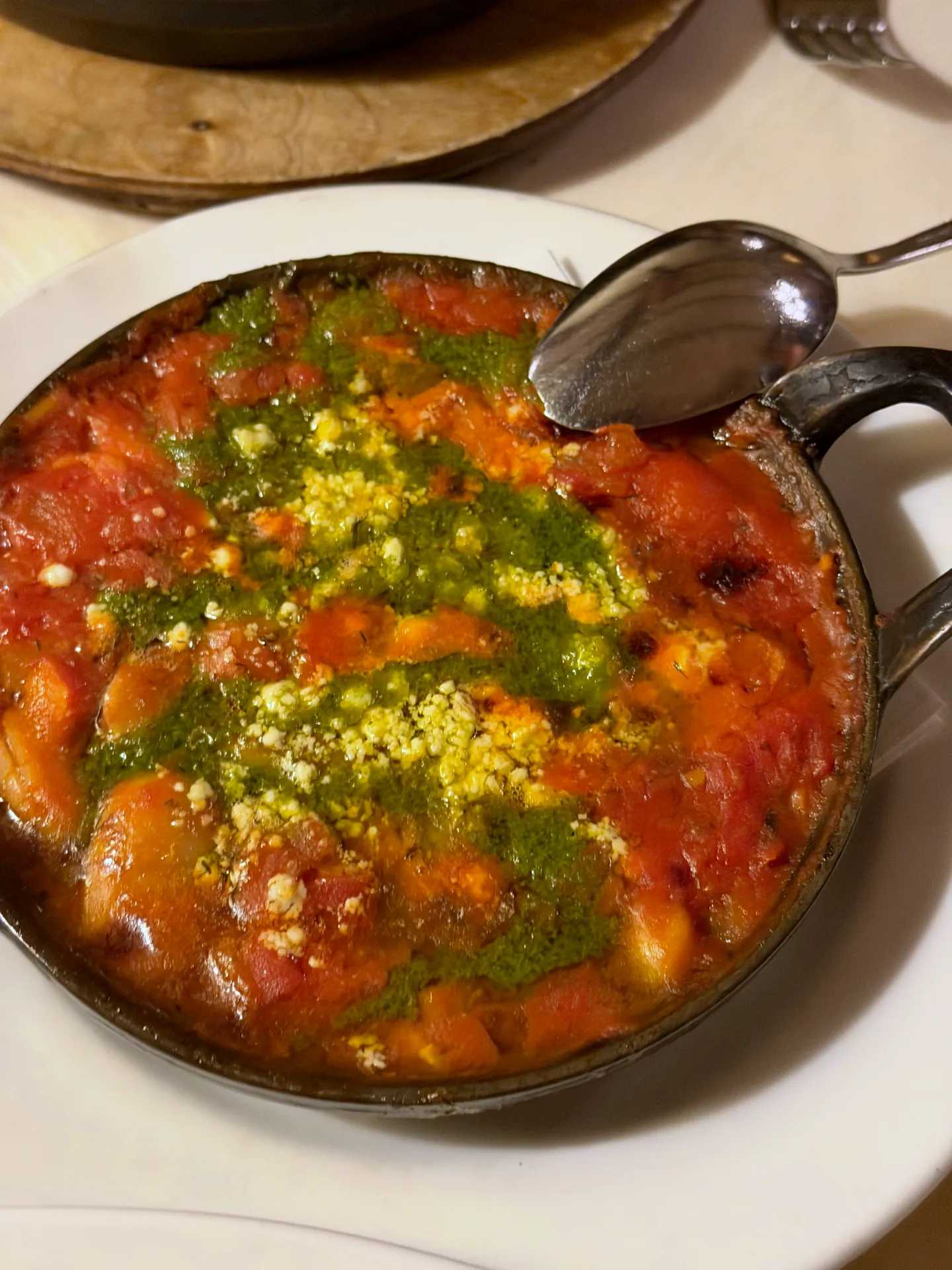 Giant baked beans in tomato sauce with herb oil and crumbled feta in a small black pan.