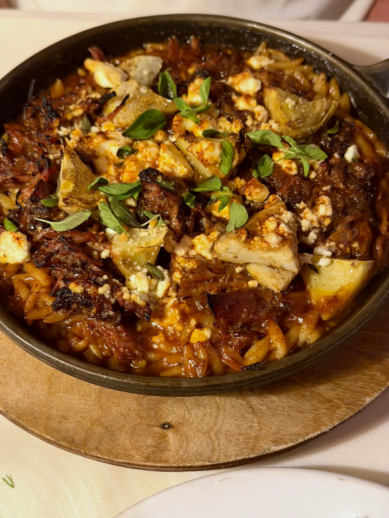 Braised goat with orzo, artichoke and feta in a black cast iron pan on a wooden board.