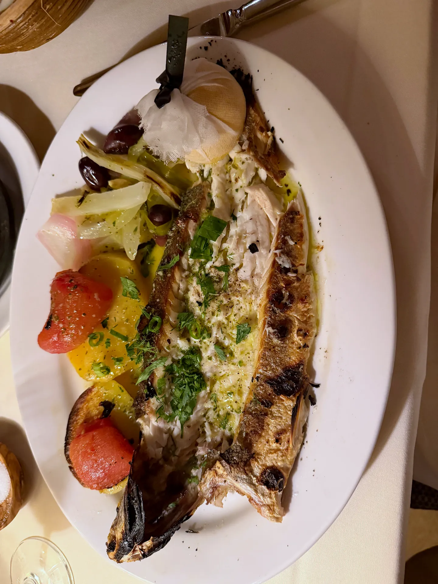 A whole grilled Mediterranean sea bass on a white plate with vegetables and herbs.
