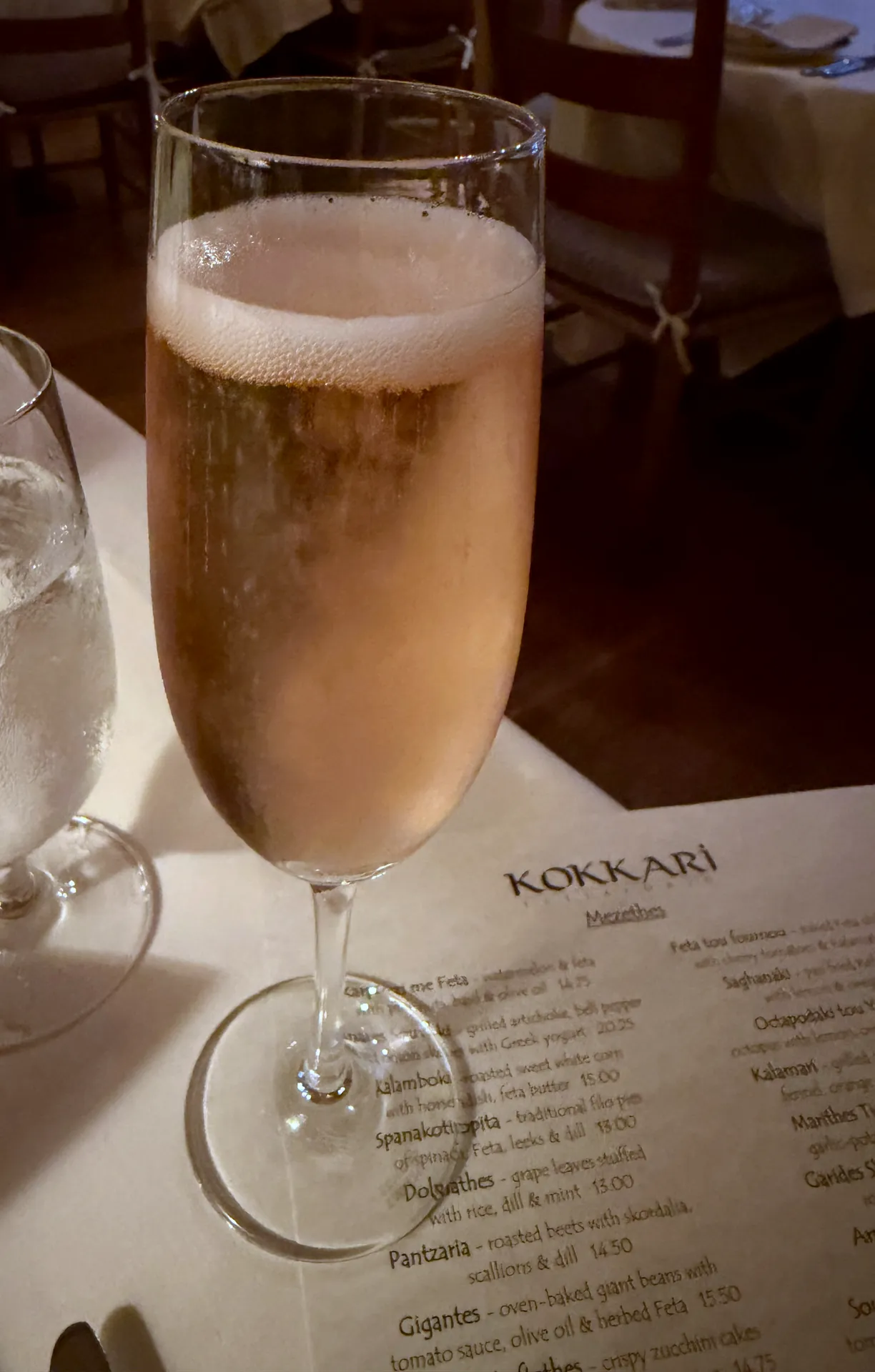 A flute of sparkling rosé standing on the Kokkari menu, with a water glass beside it.