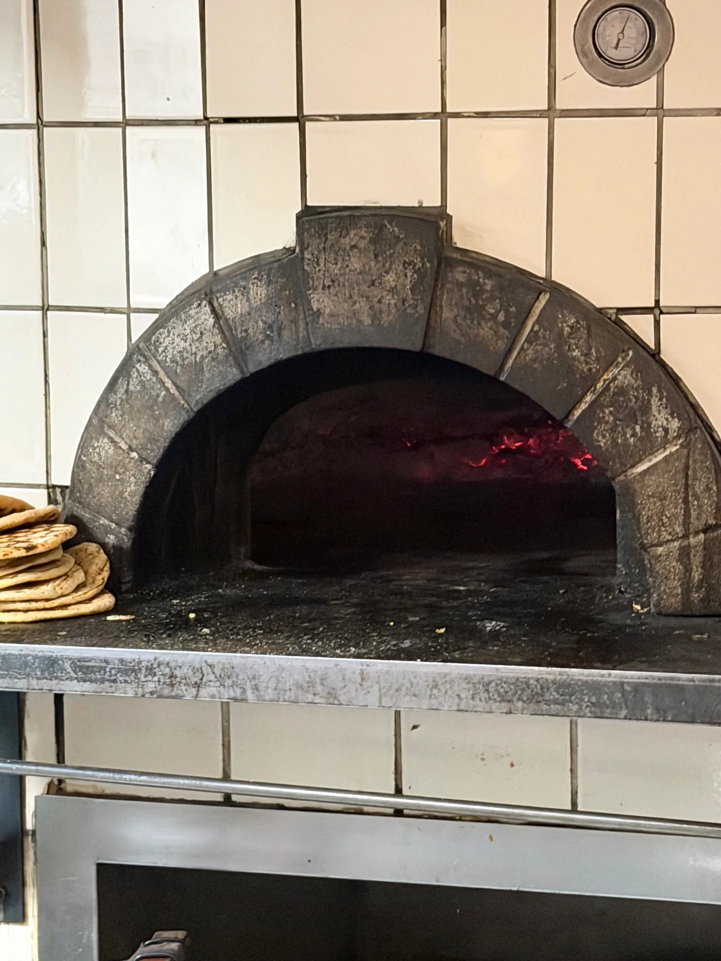 A wood-burning oven with a tiled arch and embers glowing inside, stacked flatbread beside it.