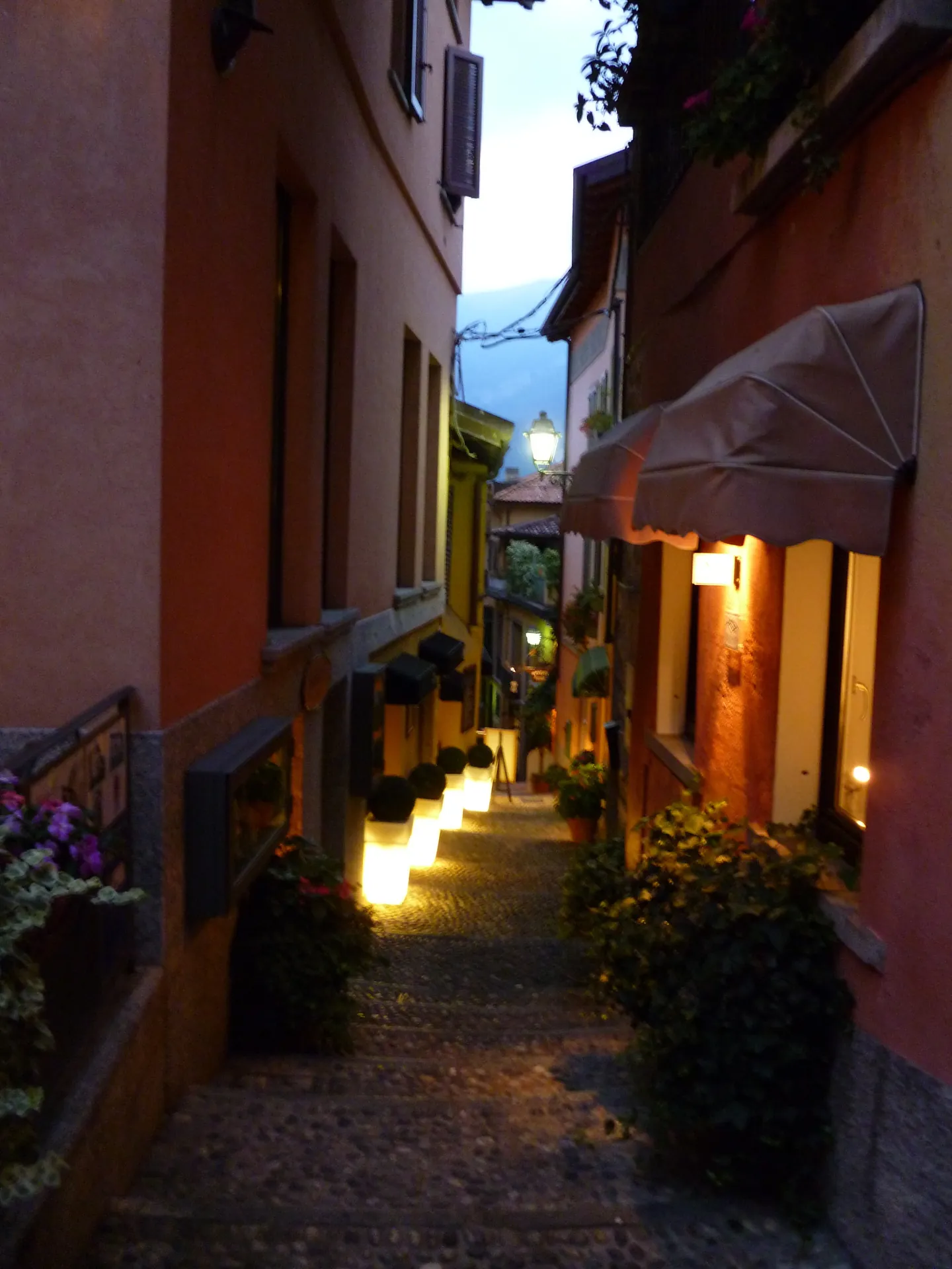 A narrow stepped alley in Bellagio at dusk, lit shopfronts and awnings on both sides.