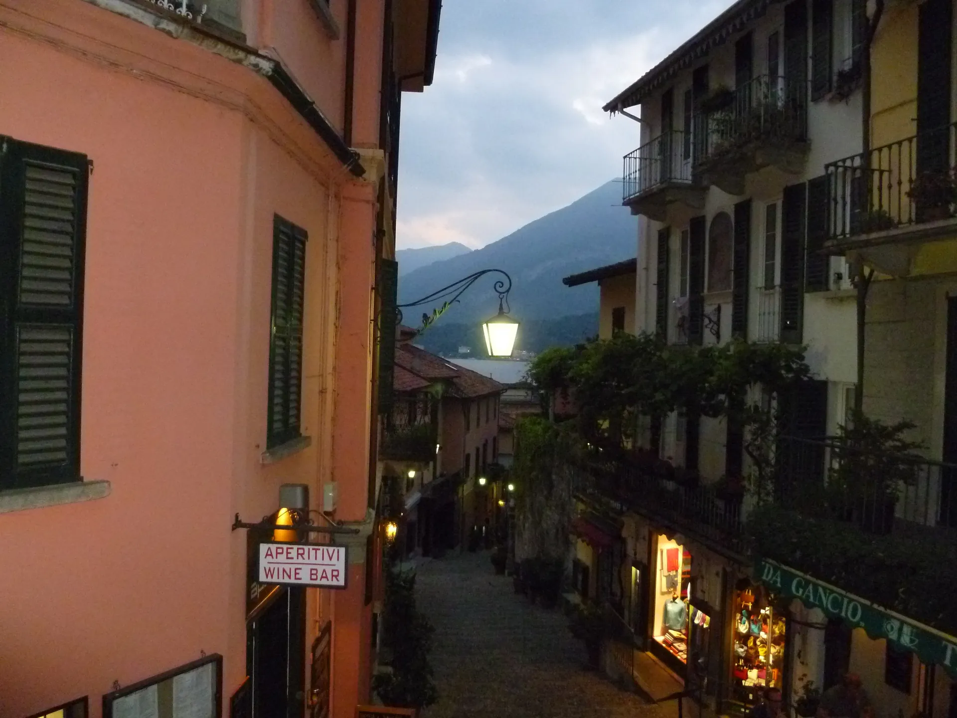 A sloping Bellagio street at dusk with a hanging lamp, opening toward the lake.