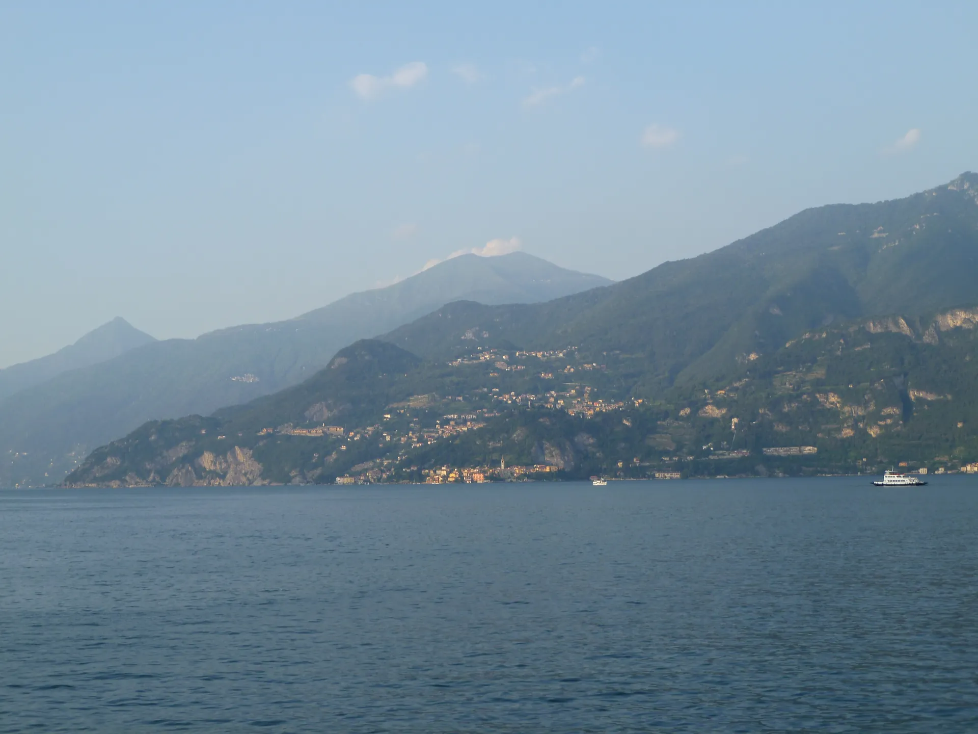 Villages on the steep western shore of Lake Como seen from the water, with a boat passing.