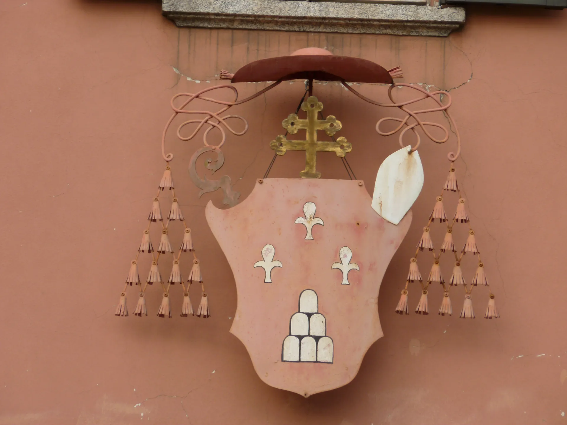 A carved coat of arms with a galero and tassels mounted on a pink wall in Lecco.