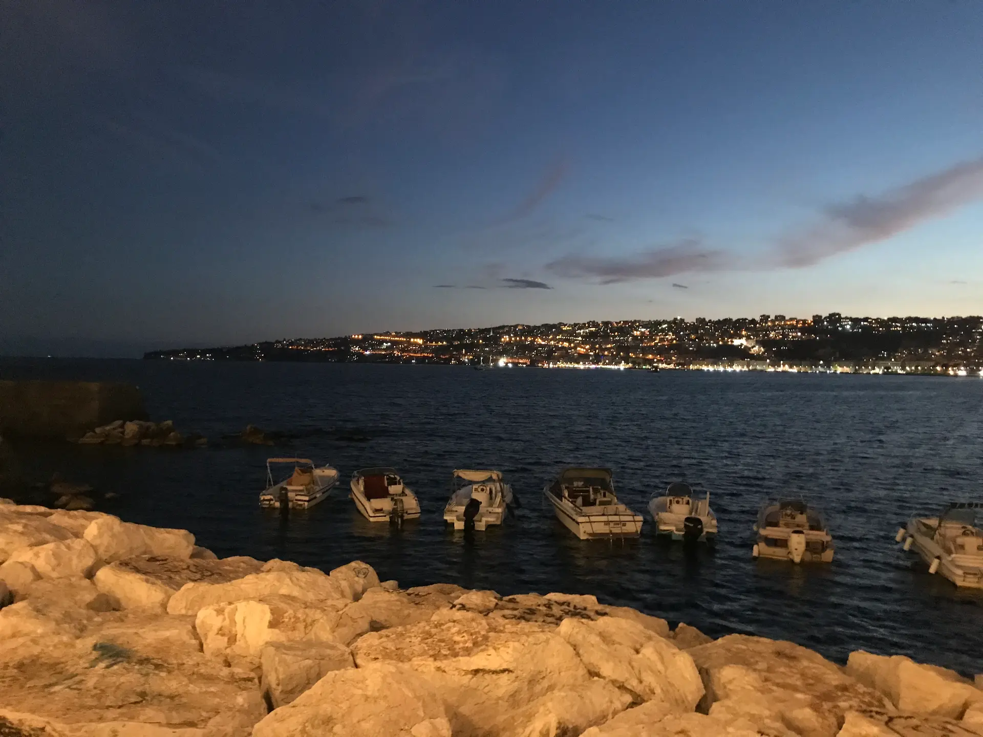 Small boats moored along the rocks of the Naples seafront at dusk.