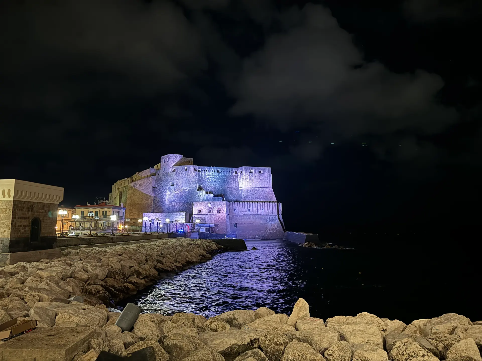 Castel dell'Ovo lit purple at night under broken cloud and a moon in Naples.