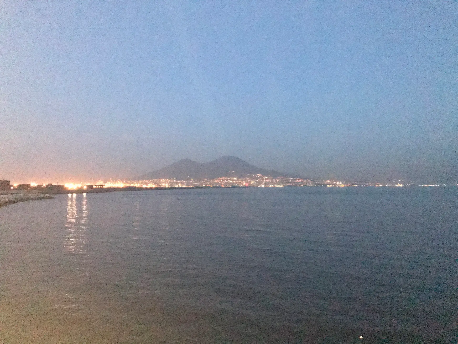 The Bay of Naples at dusk with pale sky, flat water and lights along the far shore.