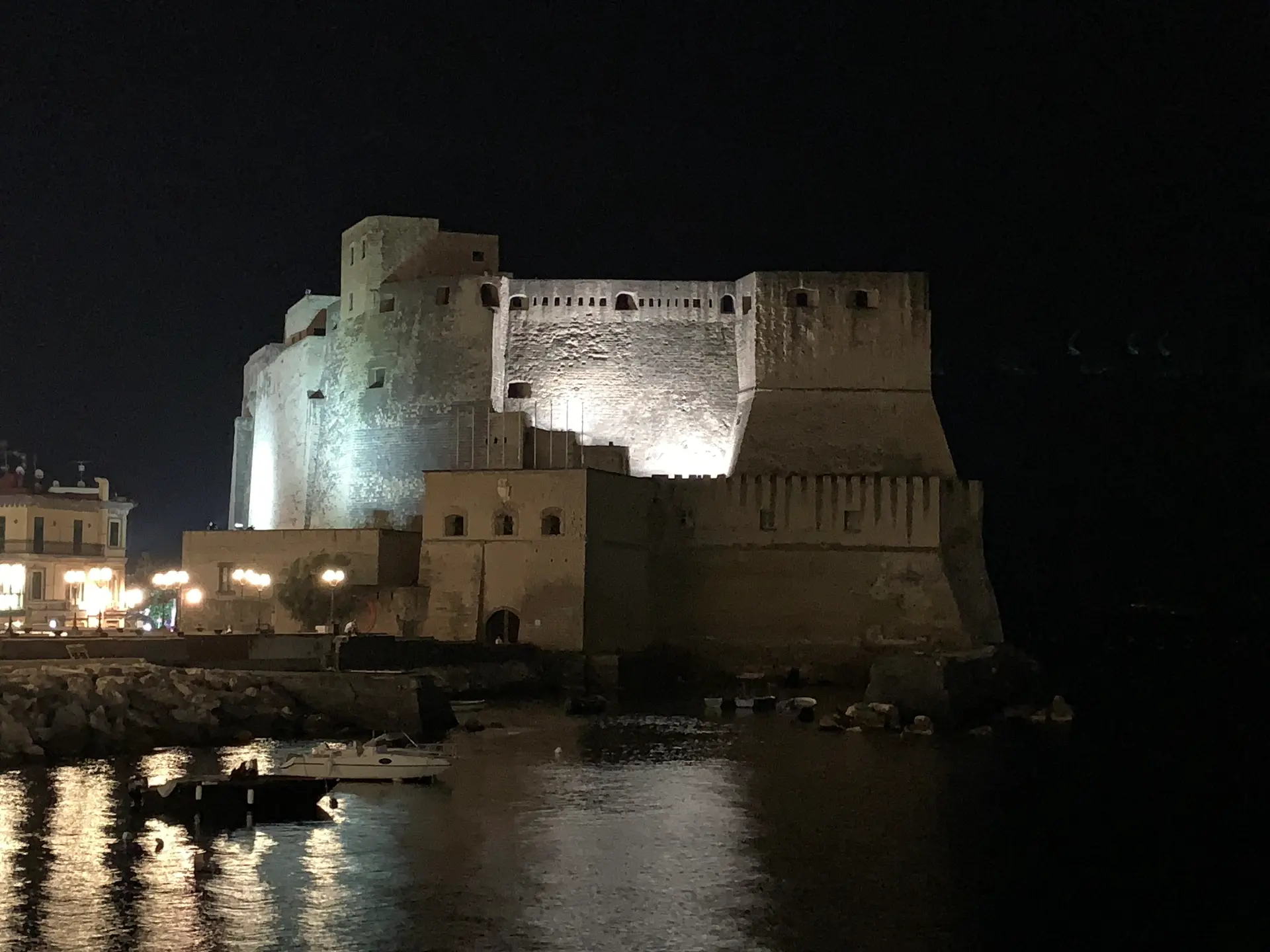 Castel dell'Ovo floodlit at night above moored boats on the Naples seafront.