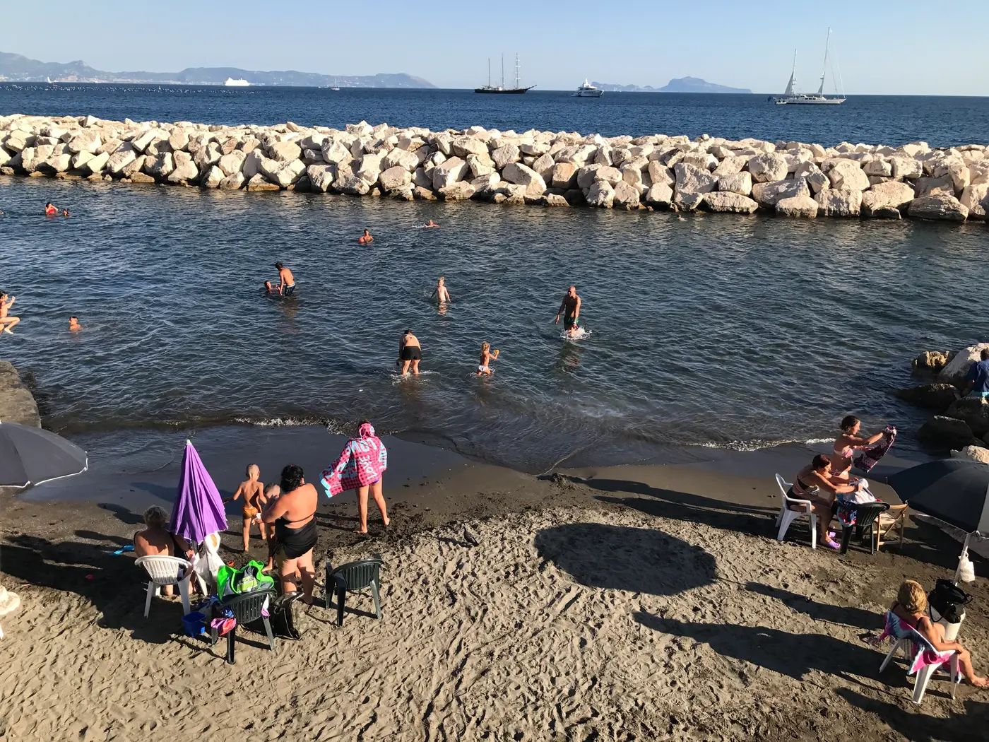 A small sand beach between breakwaters on the Naples seafront with swimmers and umbrellas.