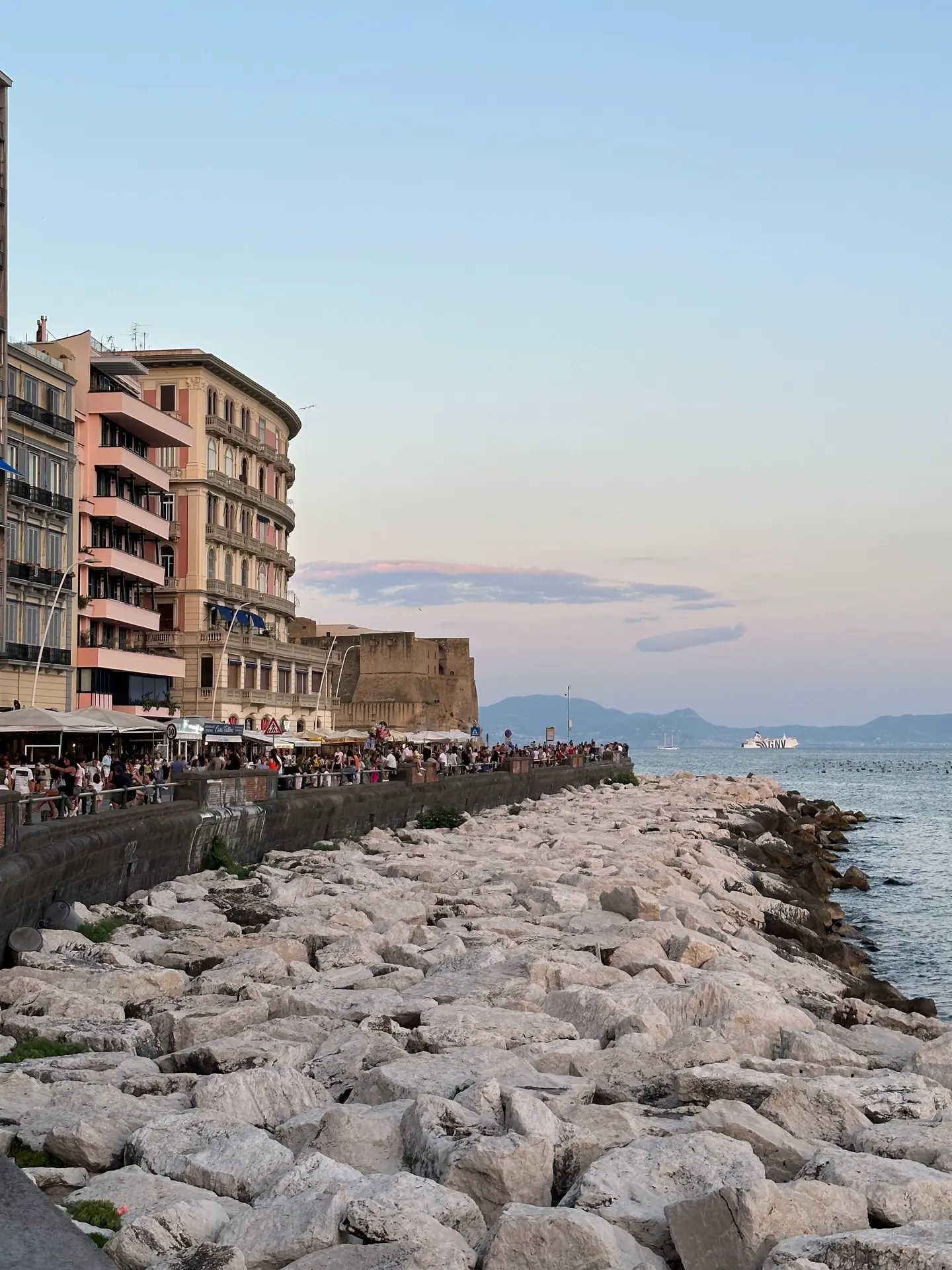 The rock breakwater and crowded hotel terraces on the Naples seafront at sunset.