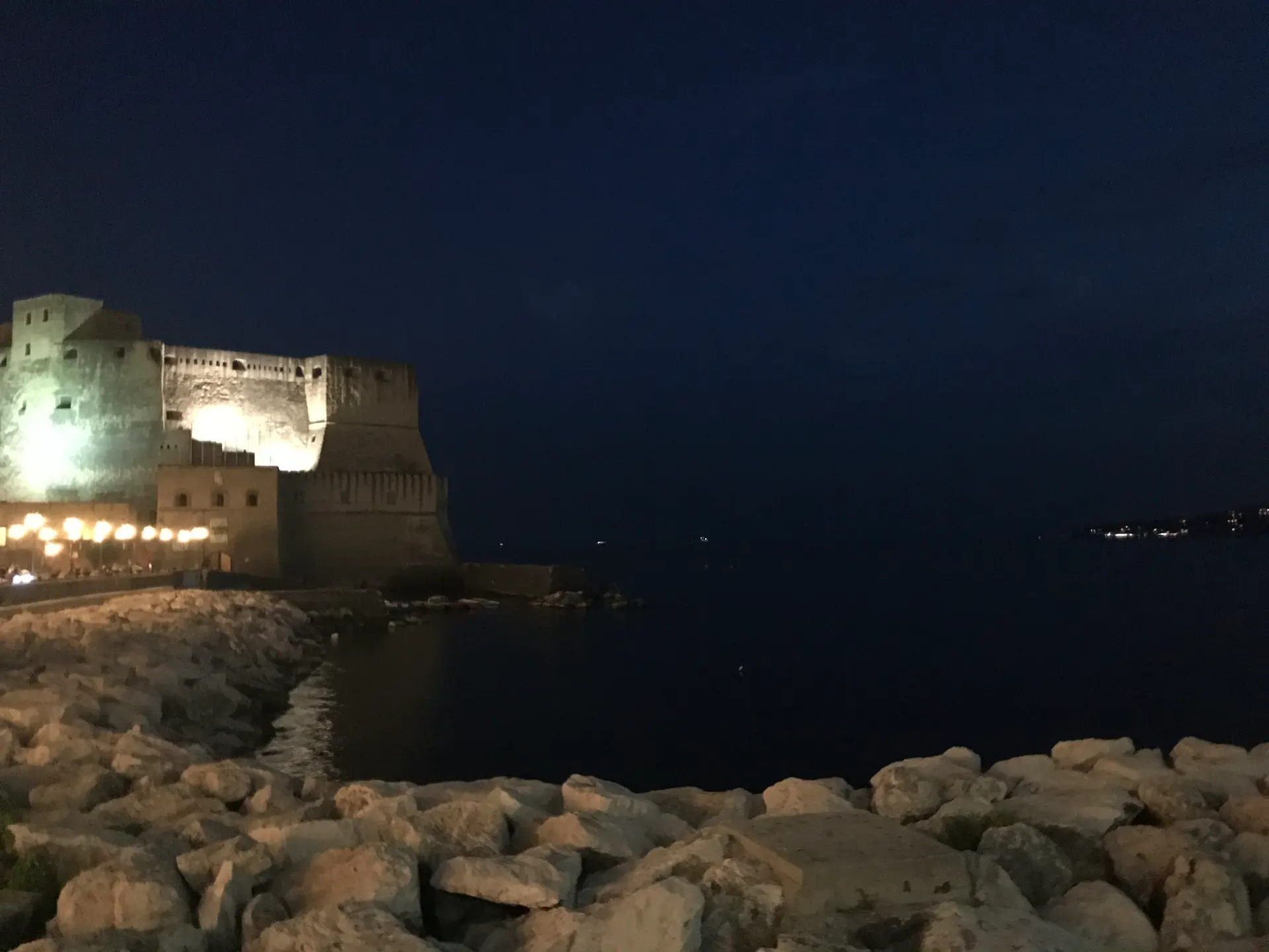 Lit restaurant terraces below the floodlit Castel dell'Ovo in Naples at night.