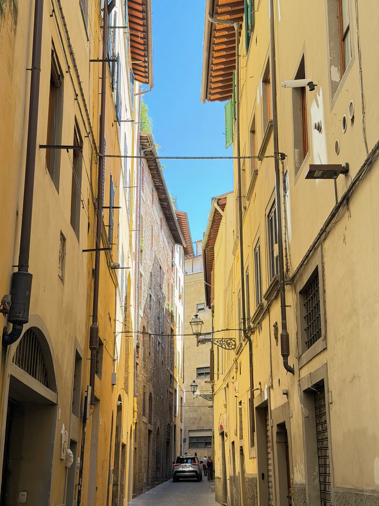 A narrow Oltrarno street in deep shade, shuttered facades on both sides and one car at the far end.