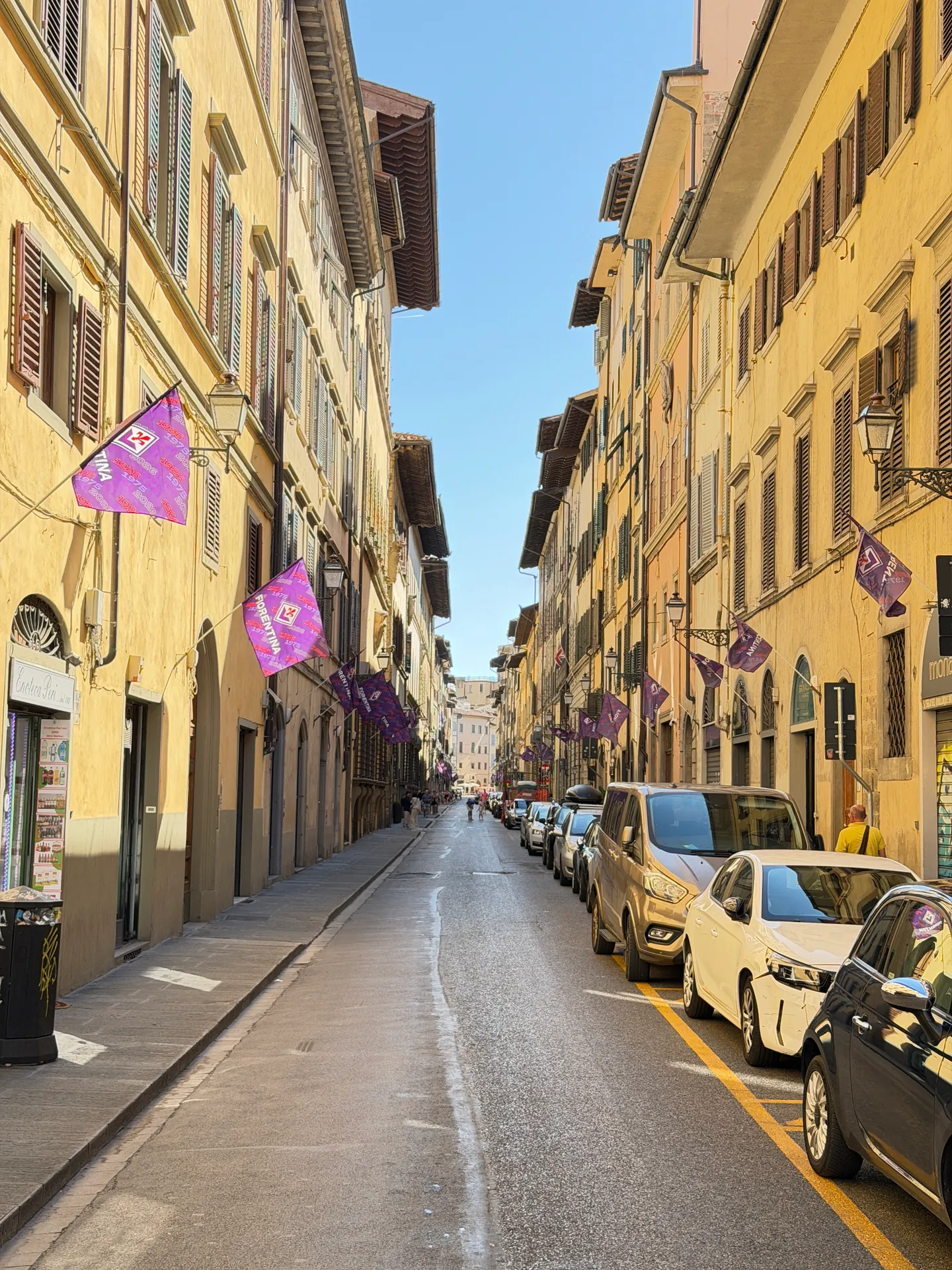 Purple pennants strung above a sunlit Oltrarno street lined with parked cars.