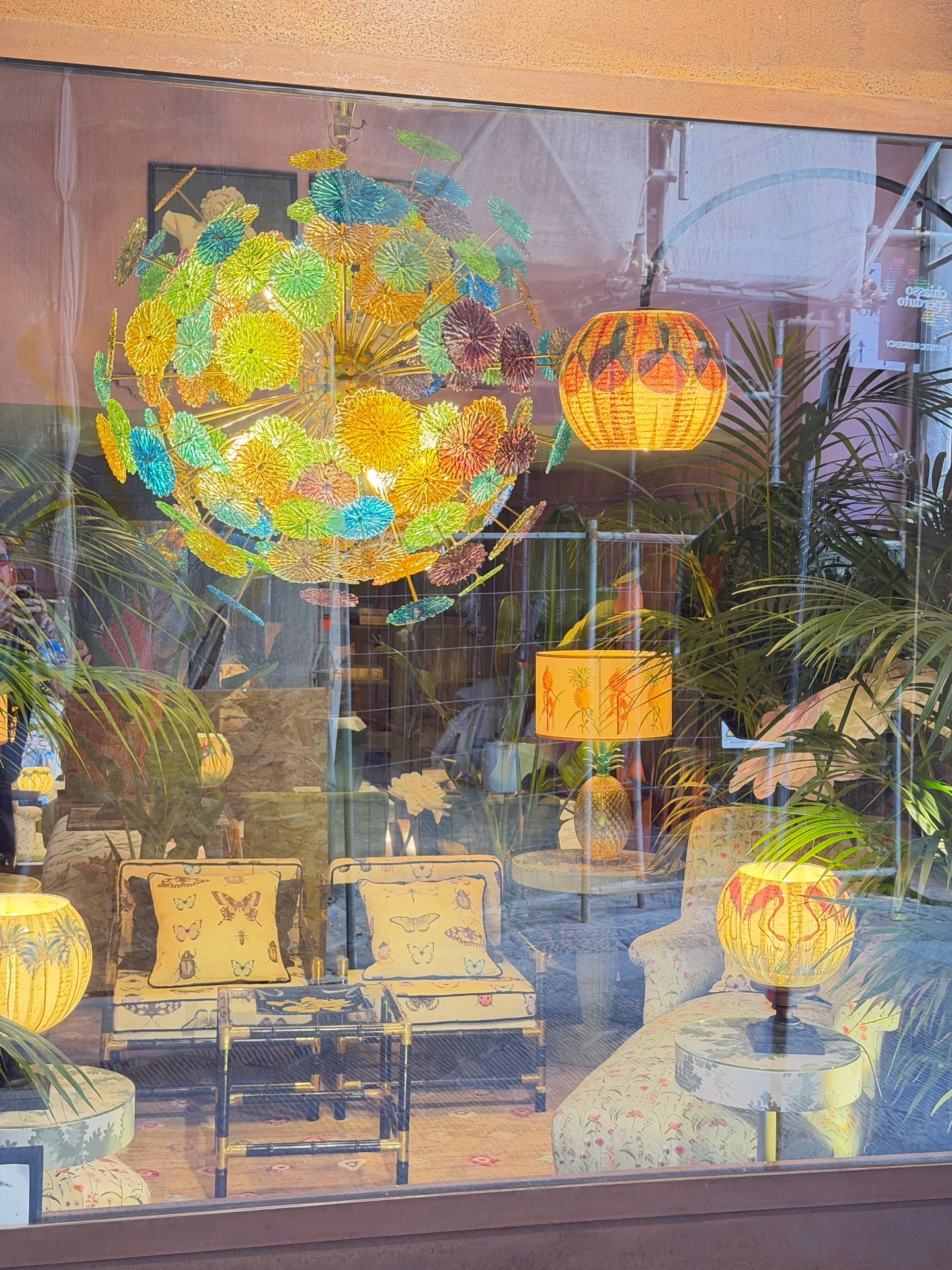 A workshop window full of coloured glass lampshades, one large globe of pierced flowers among them.