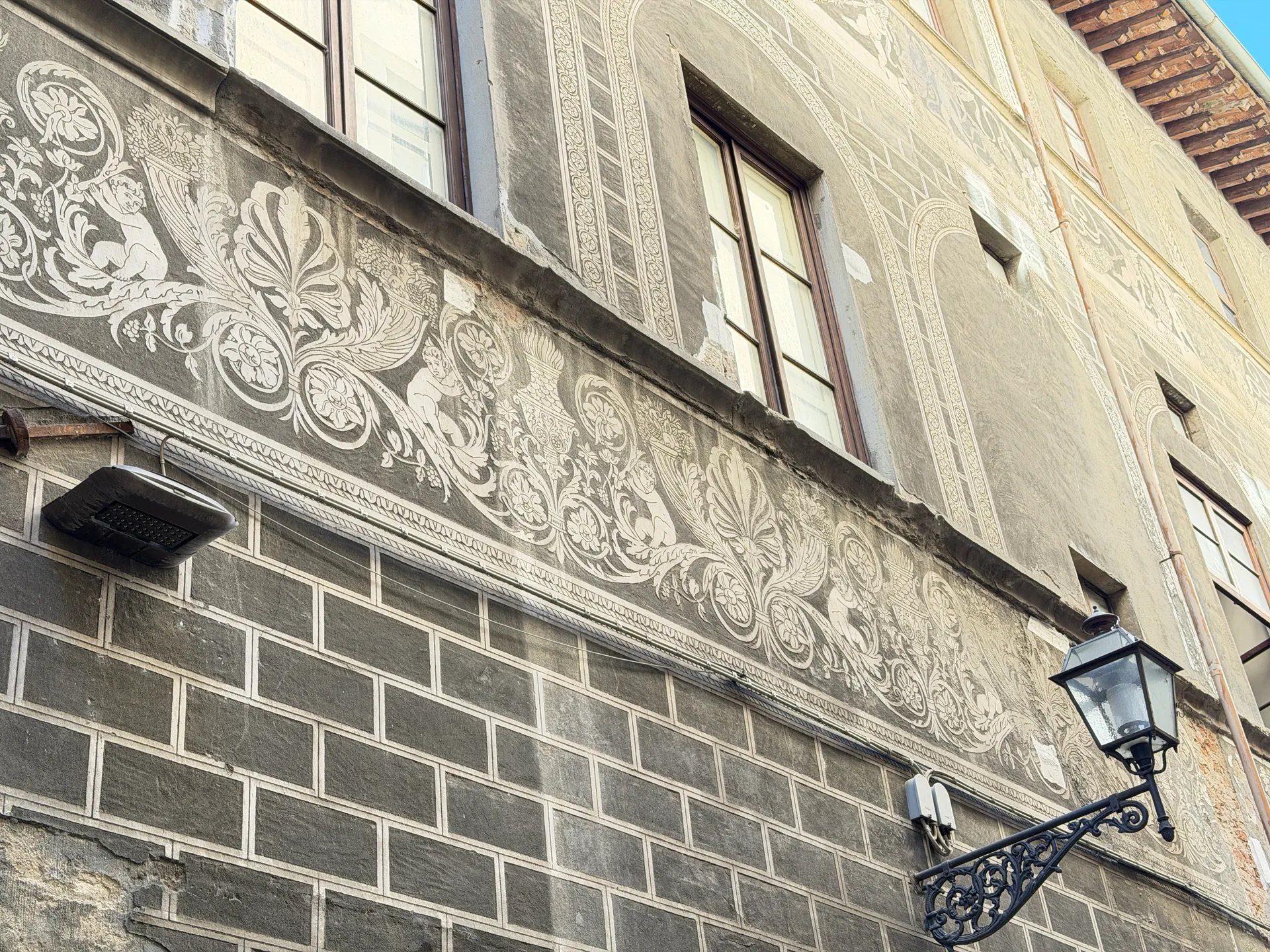 A sgraffito frieze of scrolling foliage below the windows of a rusticated palace, with an iron lamp.